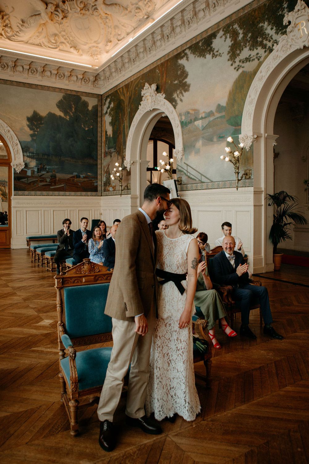 Newlyweds share a tender forehead touch as guests applaud in a grand frescoed hall.