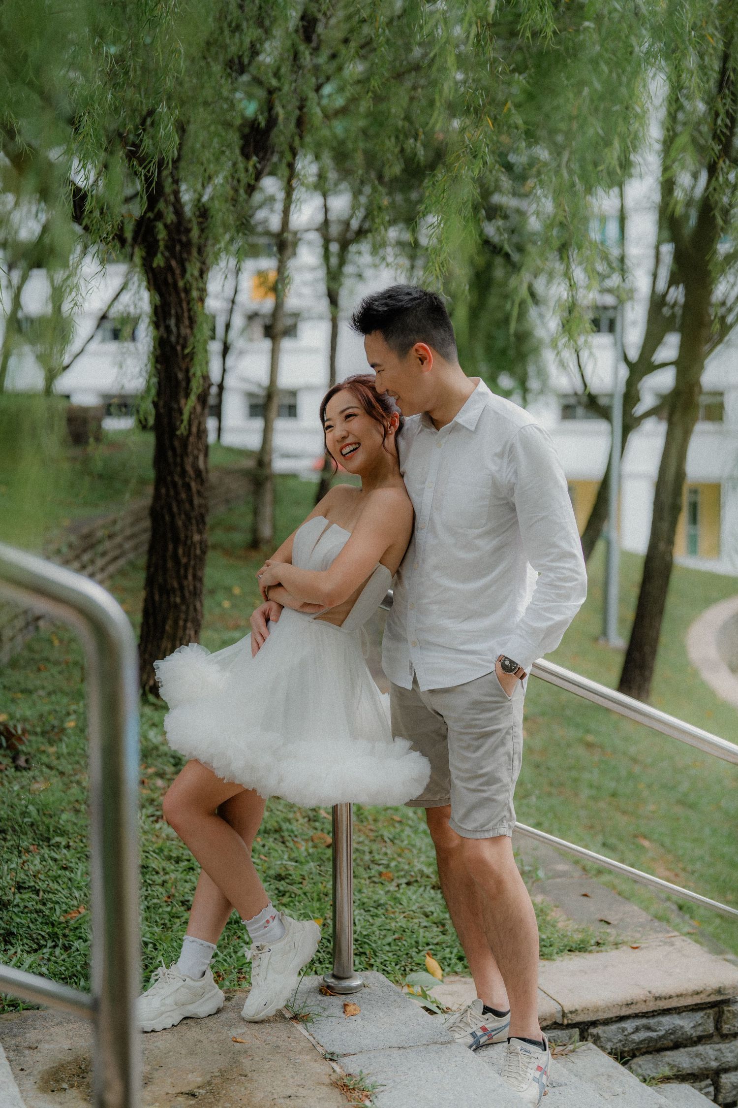 A couple poses intimately on outdoor stairs surrounded by green willow trees in a park setting.