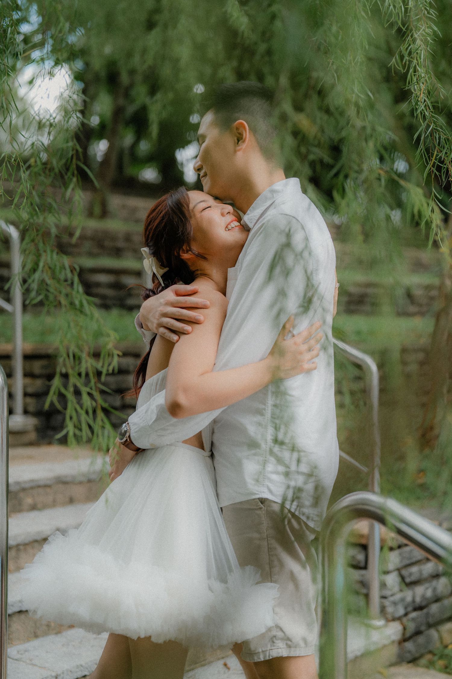 A romantic outdoor engagement photo under weeping willow trees with a couple embracing on stone steps.