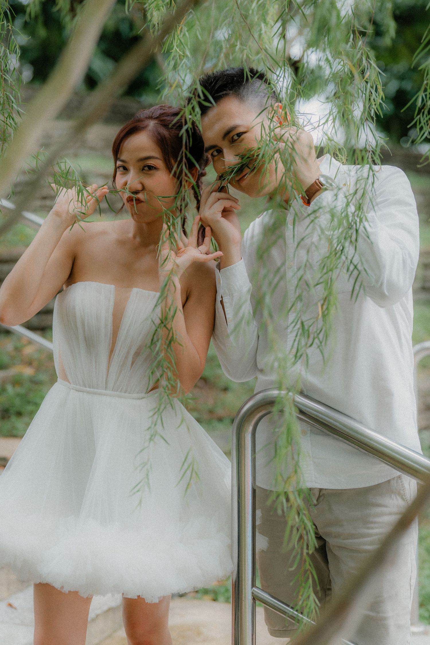 A playful moment between a couple hidden behind willow tree branches, both dressed in white outfits.