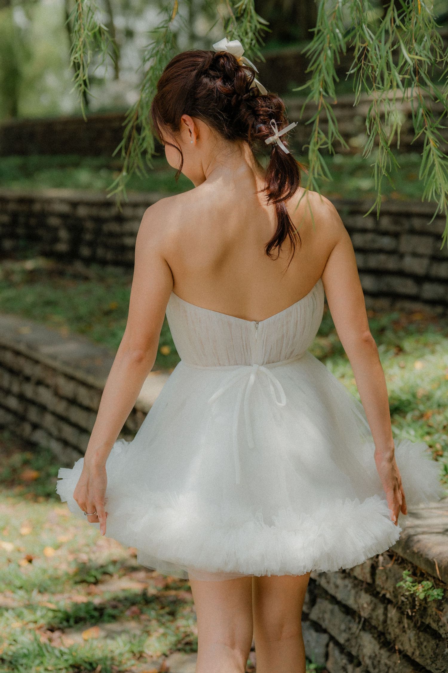 Back view of someone wearing a short white tulle dress with strapless bodice standing beneath willow branches in a garden.