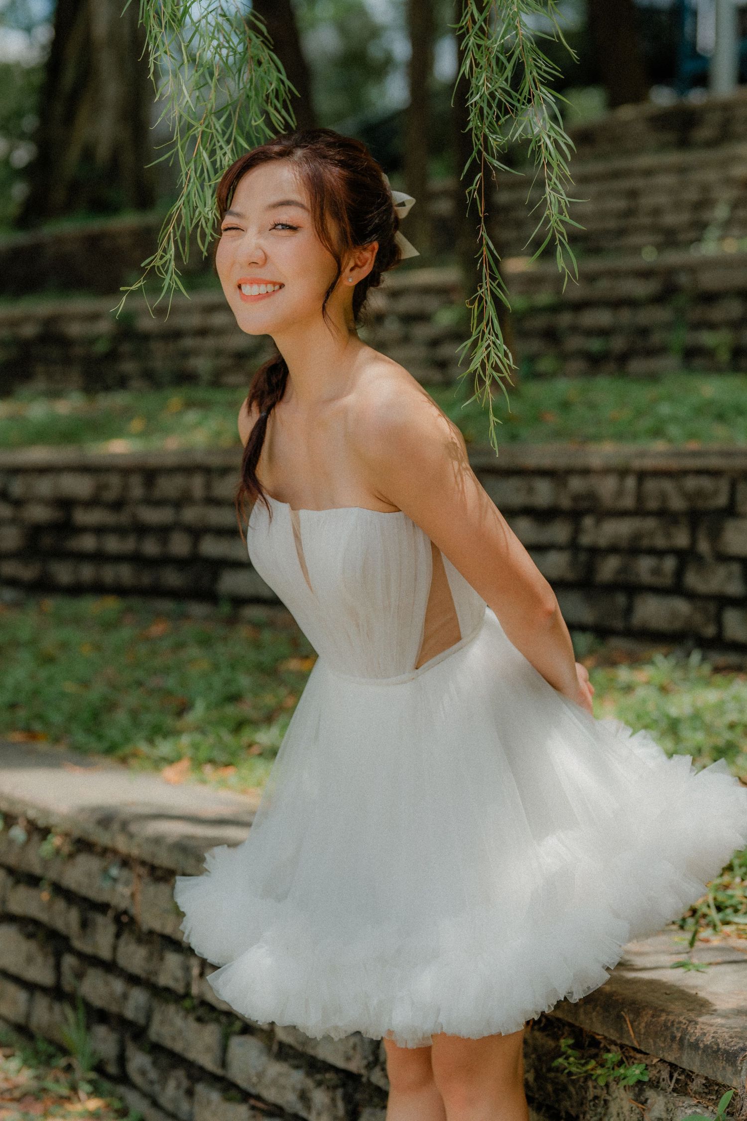 A playful shot of someone in a short white tulle dress posing by stone steps under a willow tree in a garden setting.