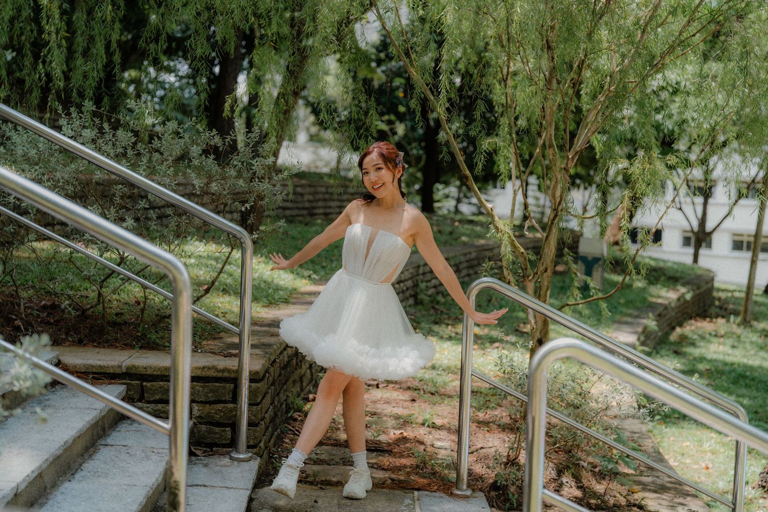 Romantic couple poses carefree in nature along garden steps with overhanging willow branches creating a dreamy scene.