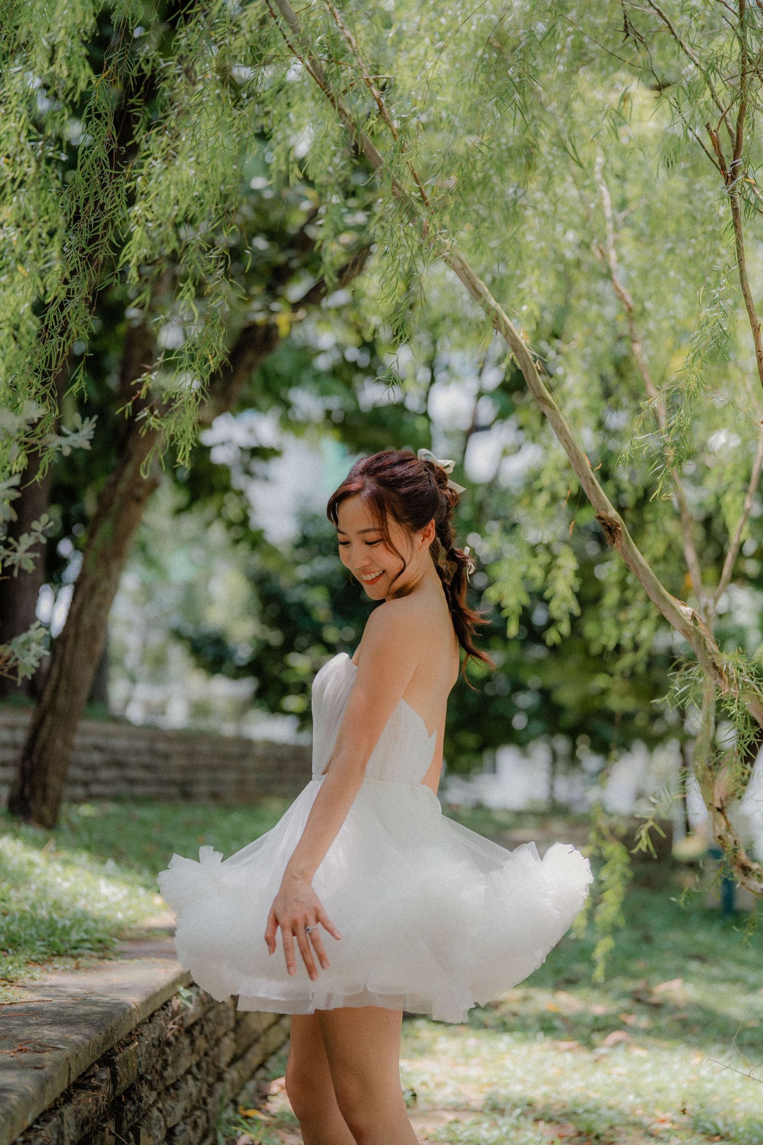 A graceful figure in a short white dress poses by a stone wall under overhanging willow branches.