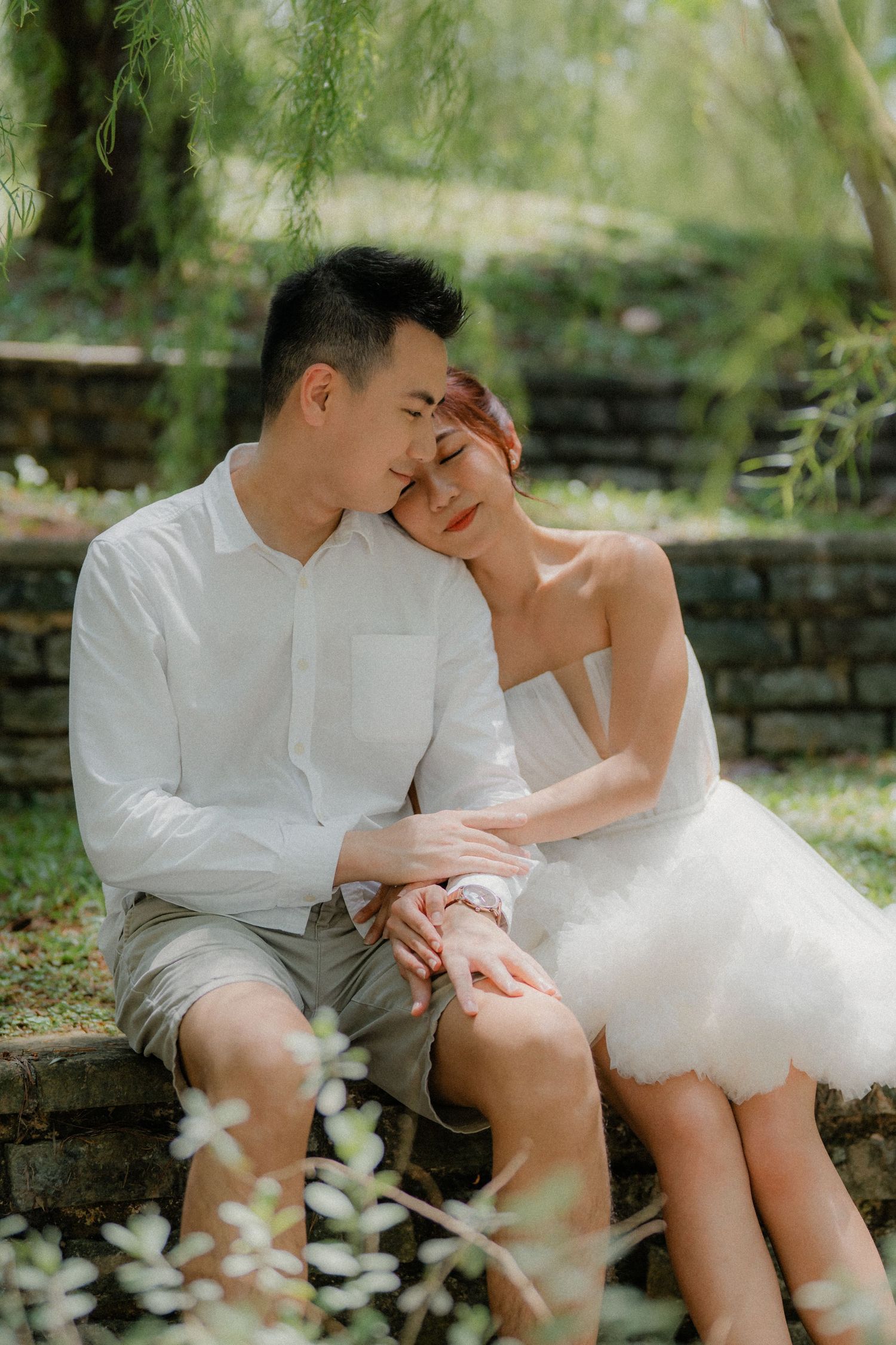 A romantic wedding photo shoot in a garden setting shows a couple sitting close together on stone steps under weeping willows.