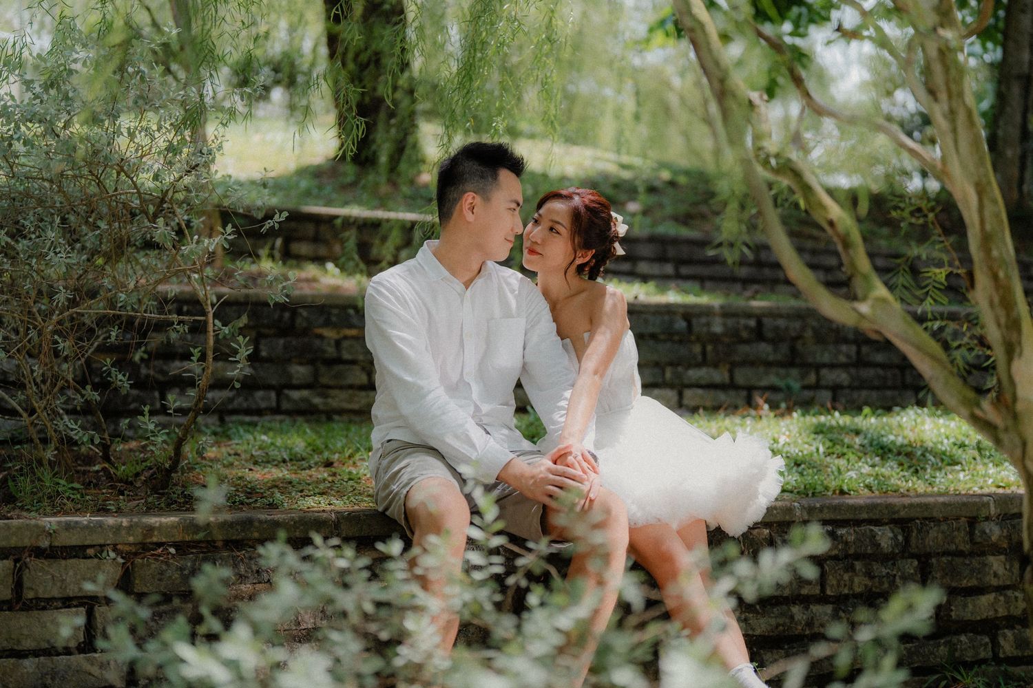 A romantic outdoor couple's wedding photo shows them sitting together on rustic stone steps surrounded by lush greenery.