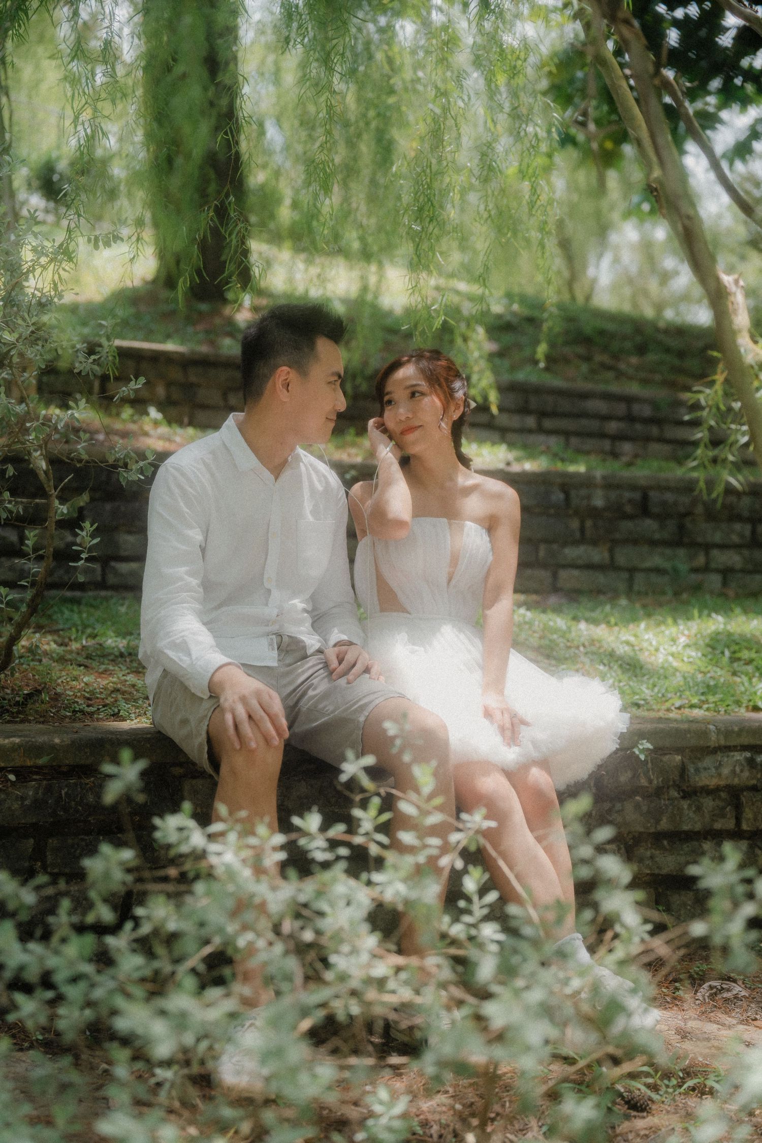 A couple in white outfits sits intimately together in nature on stone steps surrounded by greenery and willow trees.