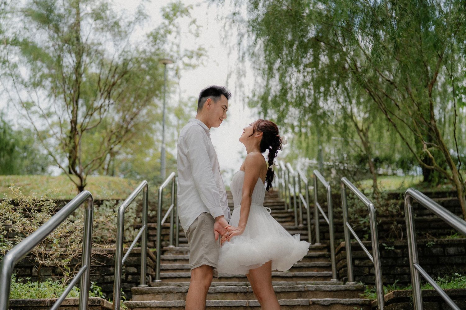A couple in wedding attire poses under weeping willow trees along a rustic staircase in a garden setting.