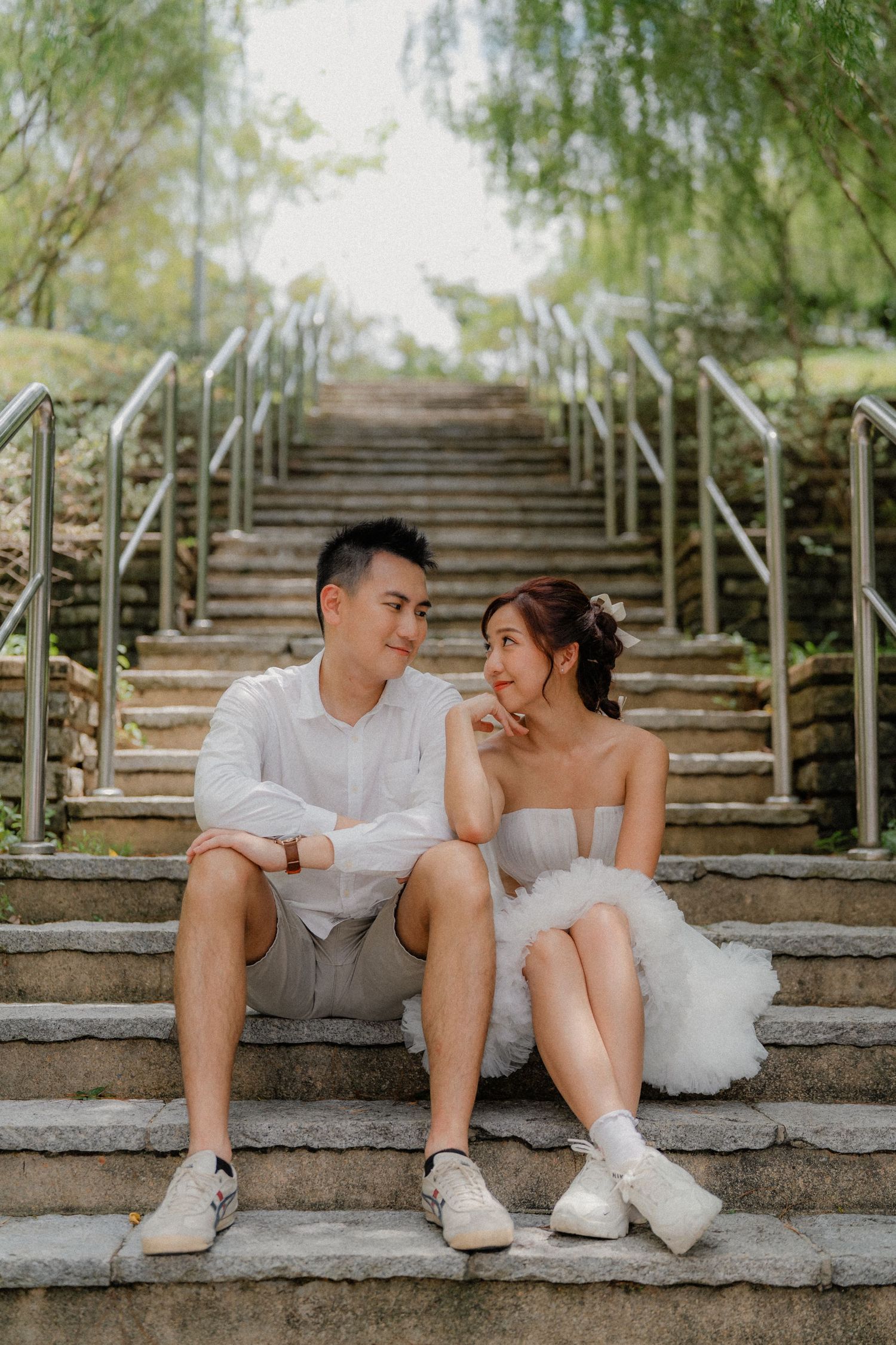A couple in white sits intimately on outdoor stone steps surrounded by lush greenery in a romantic photoshoot.