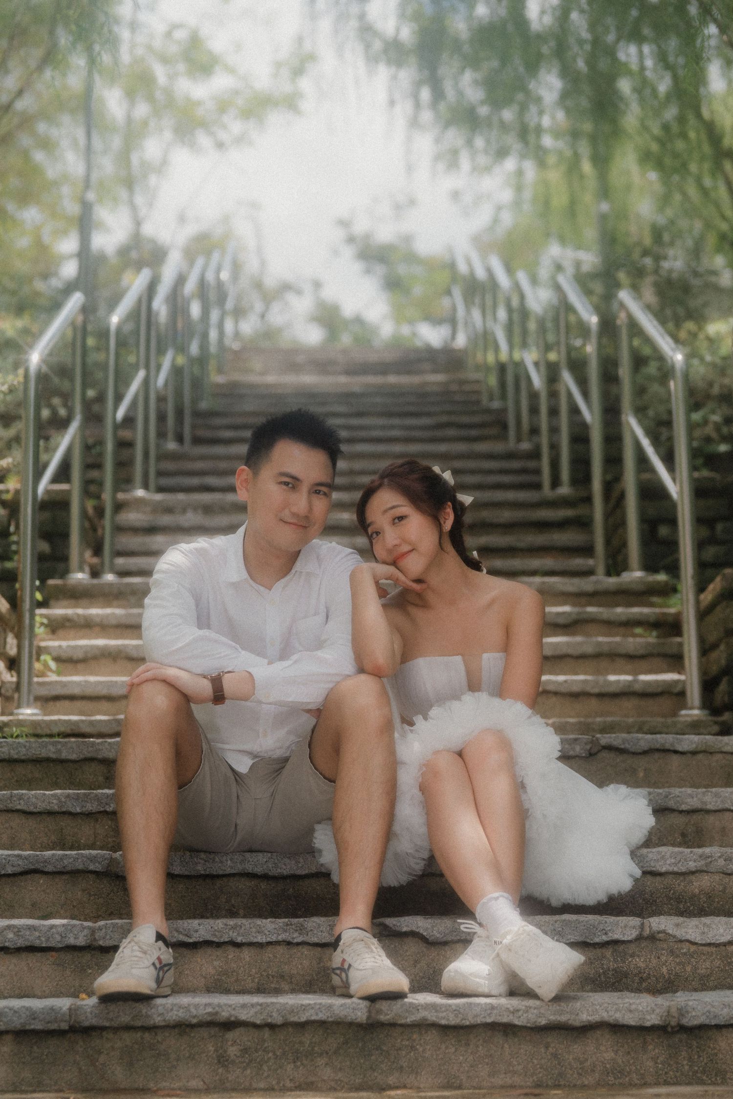 Couple in white wedding outfits share intimate moment while resting on stone staircase surrounded by trees.