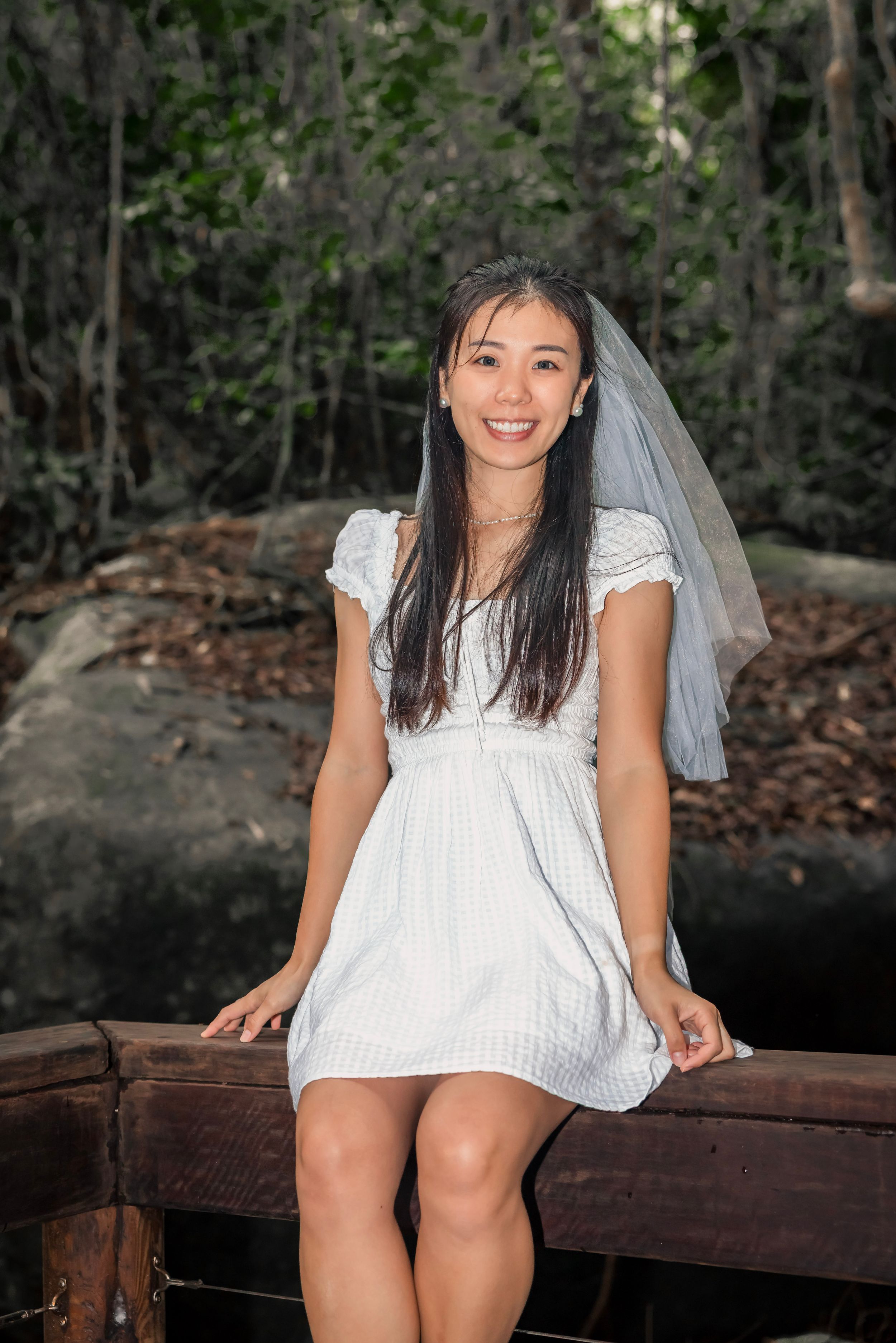 A person in a white lace dress and veil sits on a wooden bench in a forested outdoor setting.