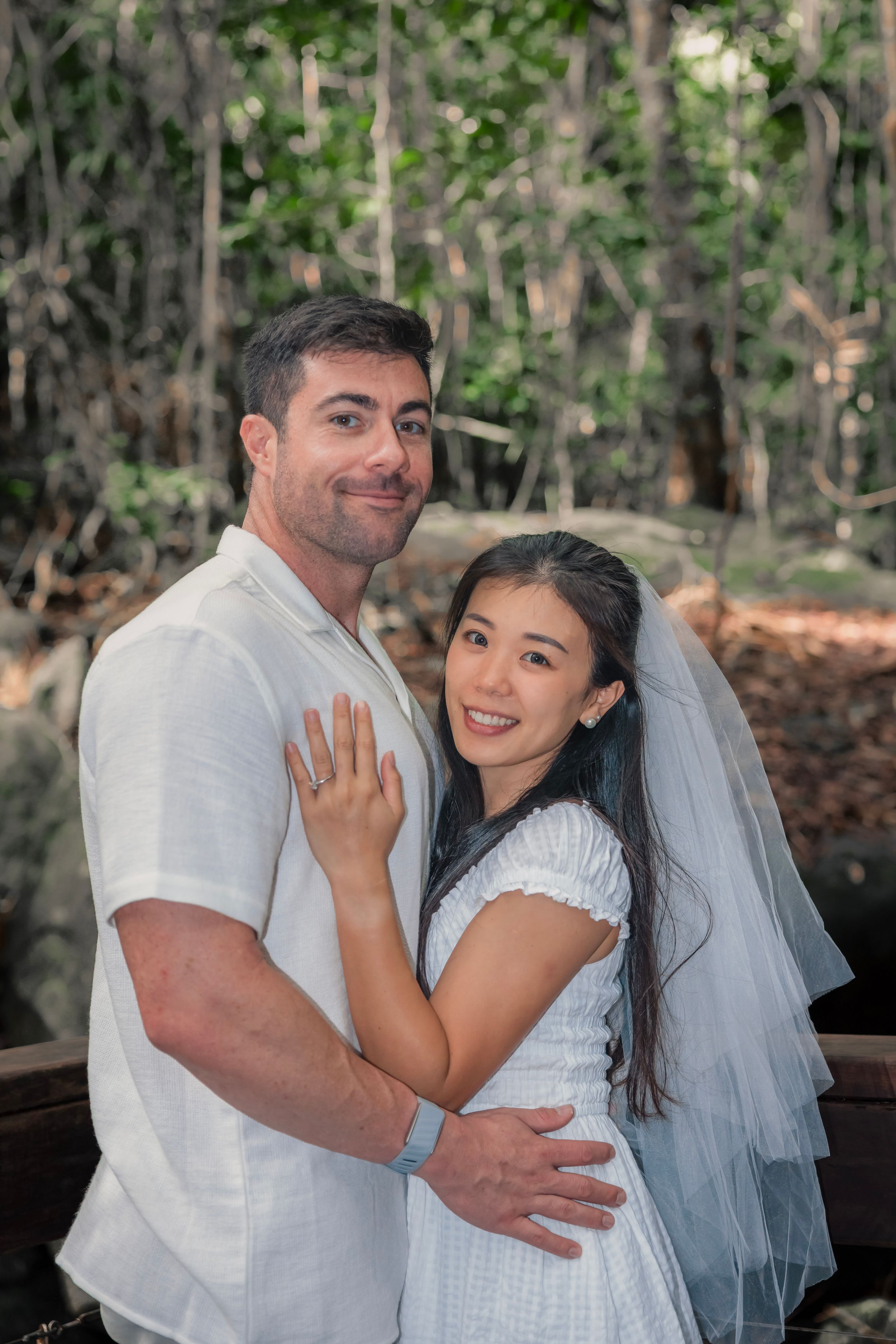 A loving newlywed couple embraces outdoors in a wooded setting while wearing white wedding attire with a flowing veil.