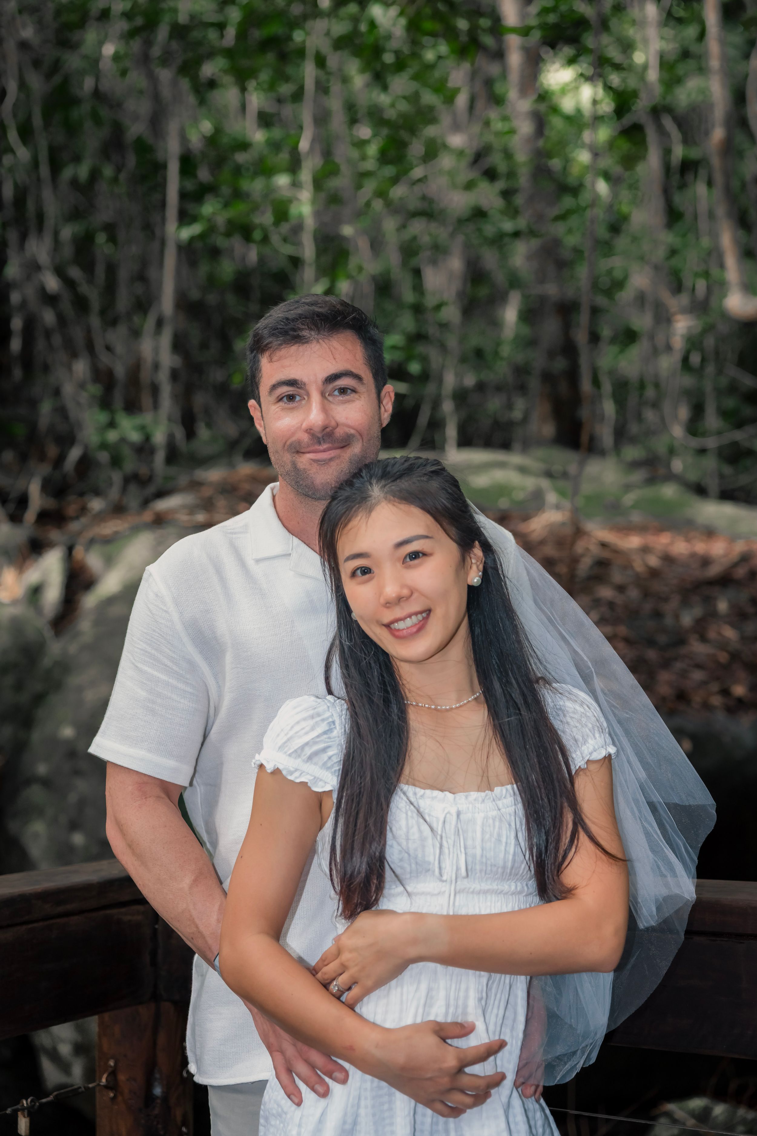 A couple embraces outdoors in white wedding attire with a forest backdrop.