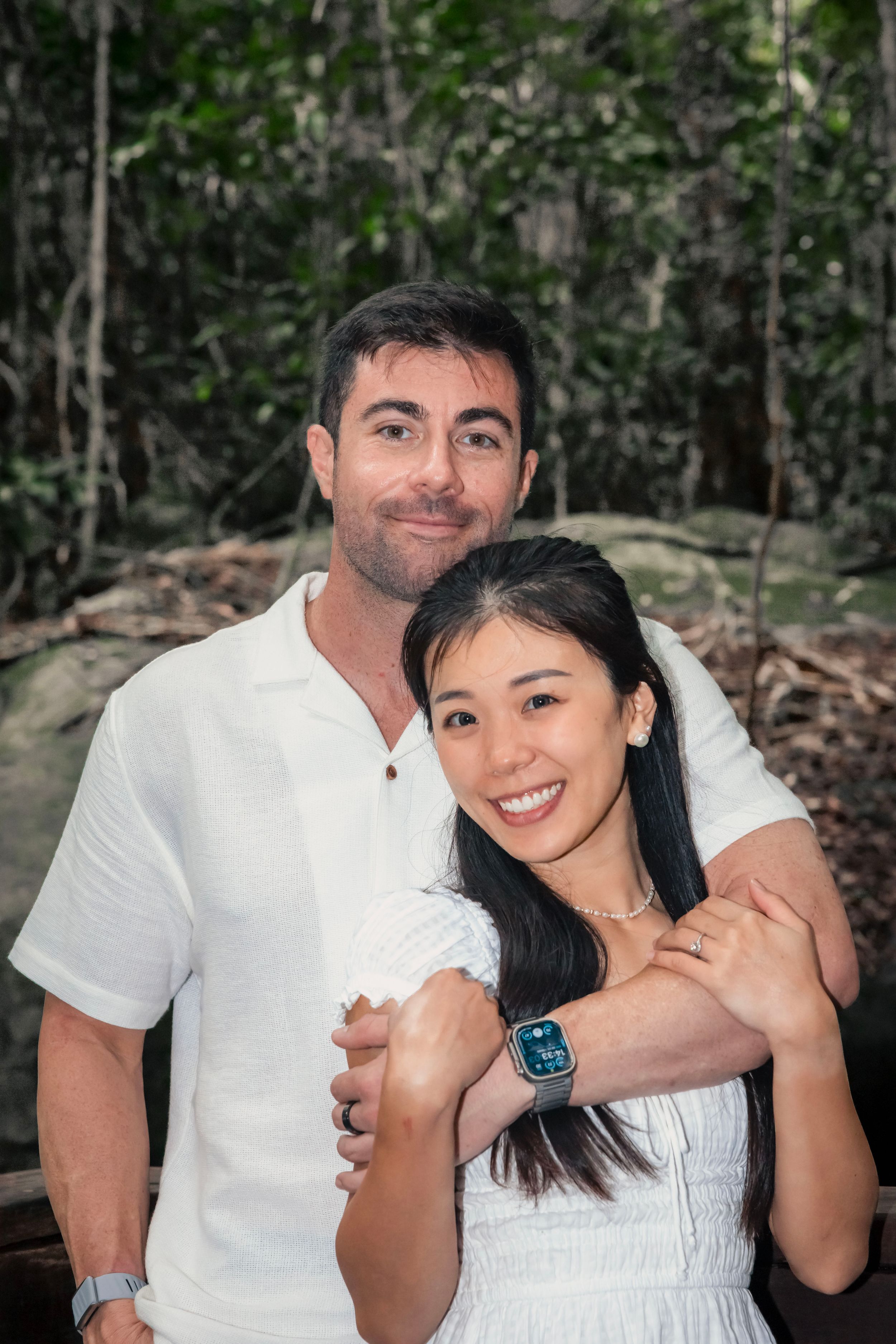A couple embraces in a natural forest setting, wearing white outfits and sharing a joyful moment together.