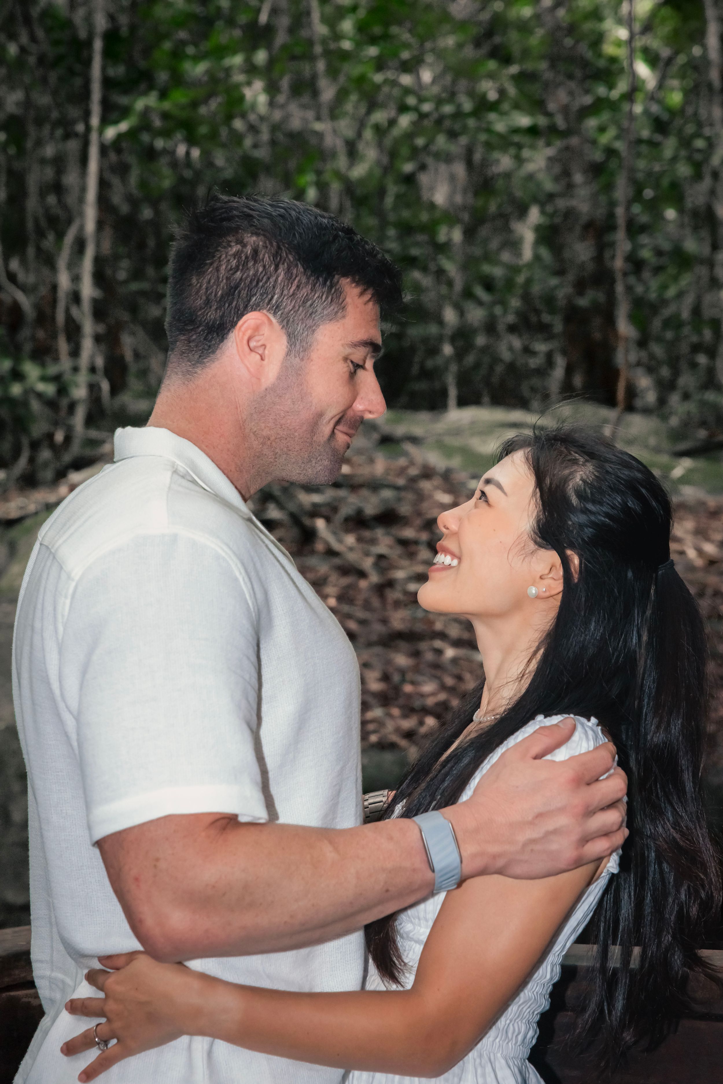 A happy couple embraces and laughs together while posing for romantic photos in a wooded outdoor setting.