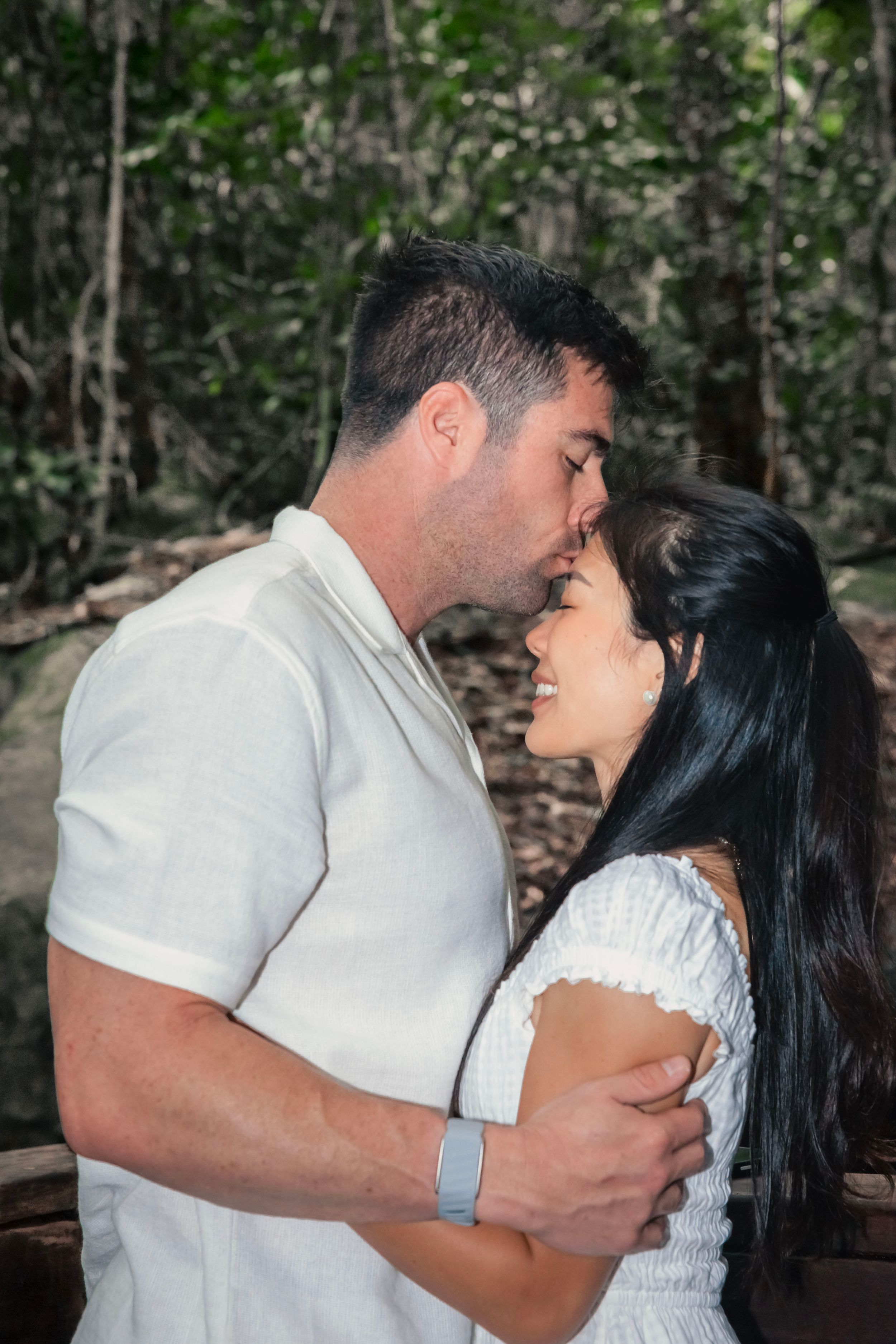 A romantic couple embraces tenderly in a forest setting, both wearing white as they share an intimate moment together.