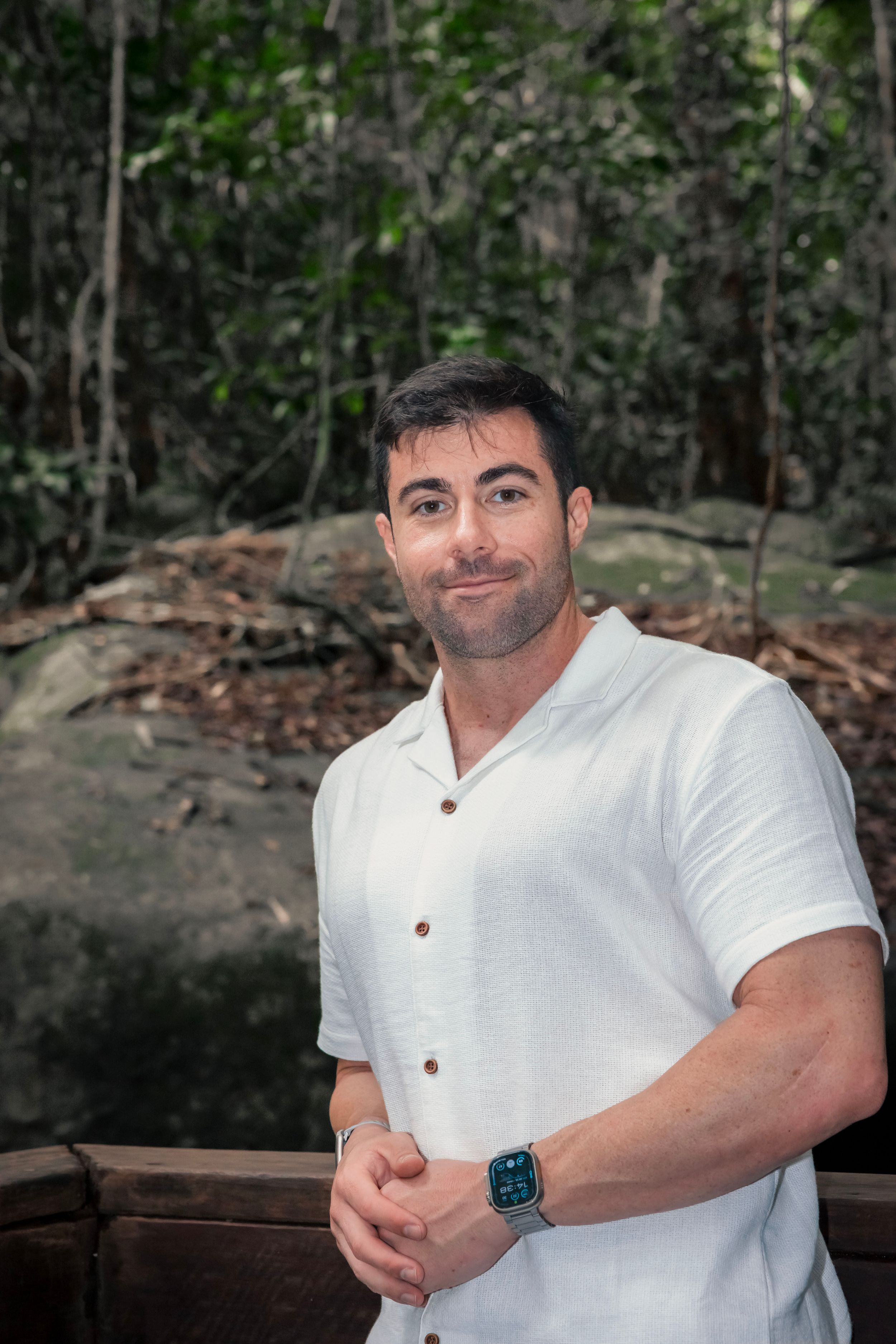 Person in white henley shirt poses against wooden railing outdoors in a forest setting.
