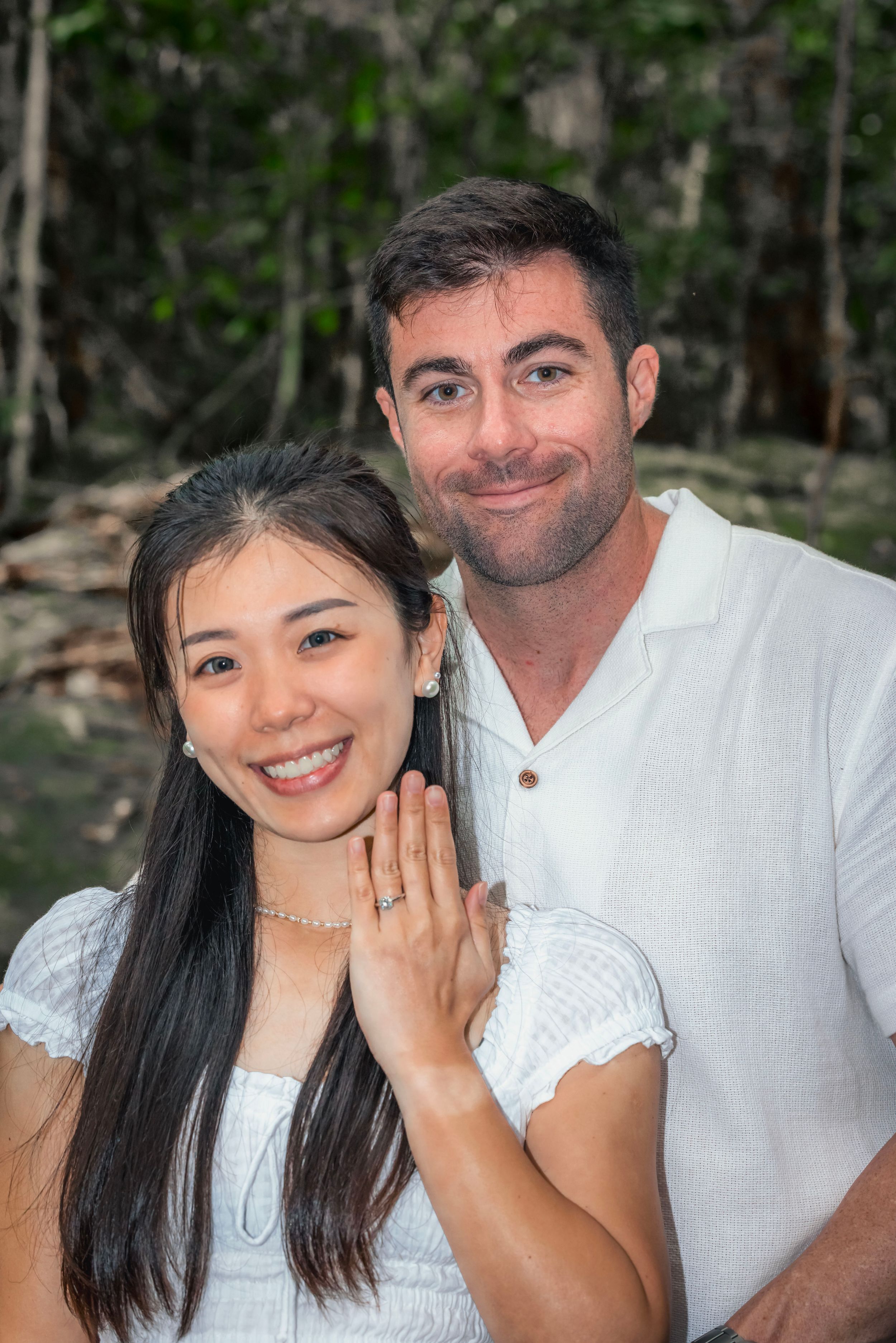 A couple smiles together outdoors while showing off an engagement ring on her hand.