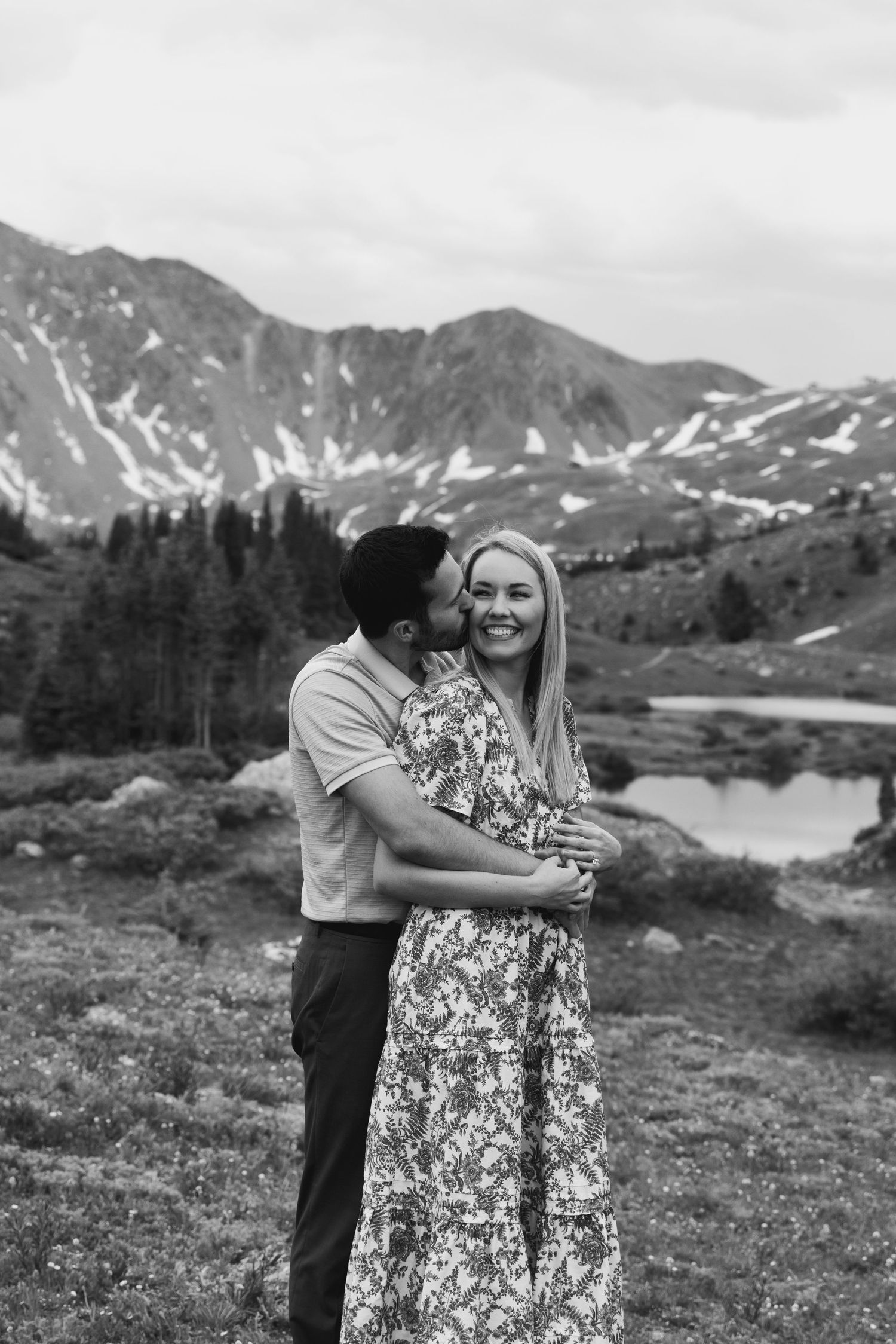 A couple embraces in front of a scenic mountain landscape with snowy peaks and a lake in the background at Loveland Pass, Colorado.