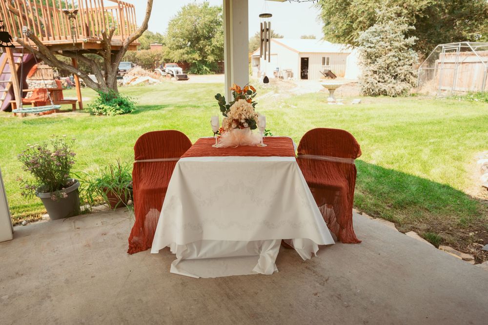A rustic wedding reception table with two red chairs set up on a covered patio overlooking a grassy lawn.