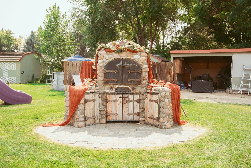 A rustic stone wedding arch decorated with burnt orange fabric stands in a backyard setting.
