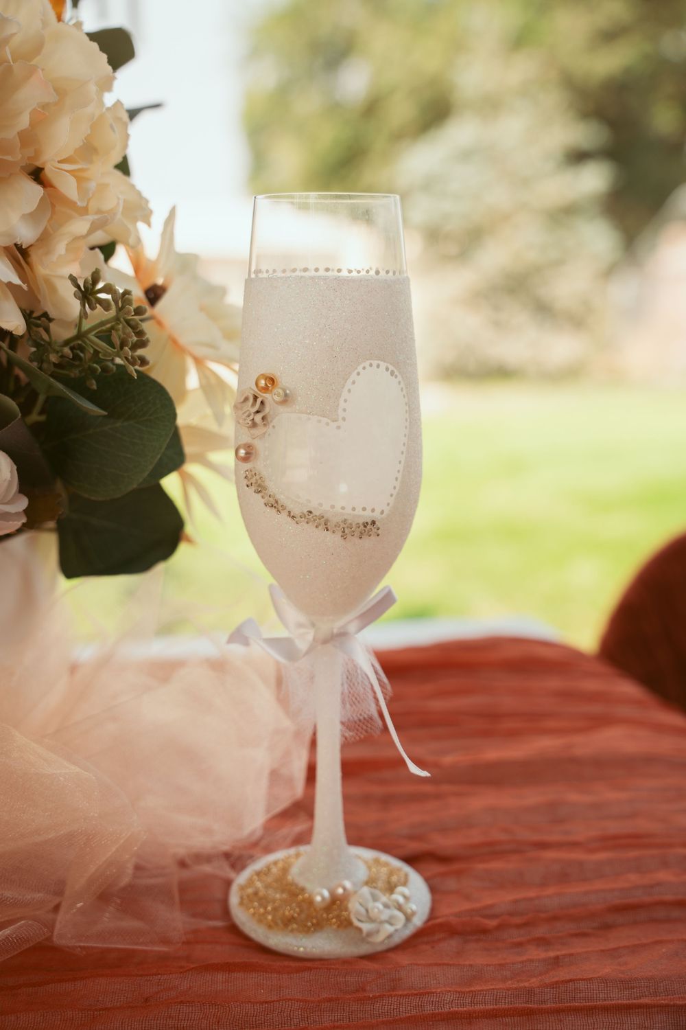 A decorative wine glass adorned with delicate lace and tulle details sits on a rust-colored tablecloth.