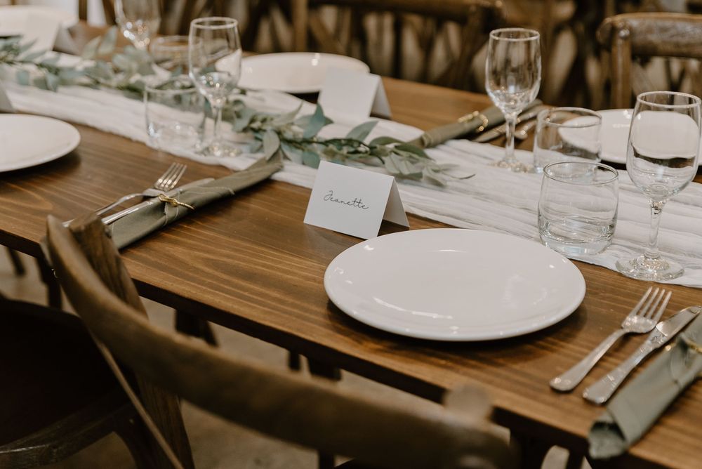 Elegant place setting at wedding reception with wooden table, white plates, wine glasses and eucalyptus greenery.