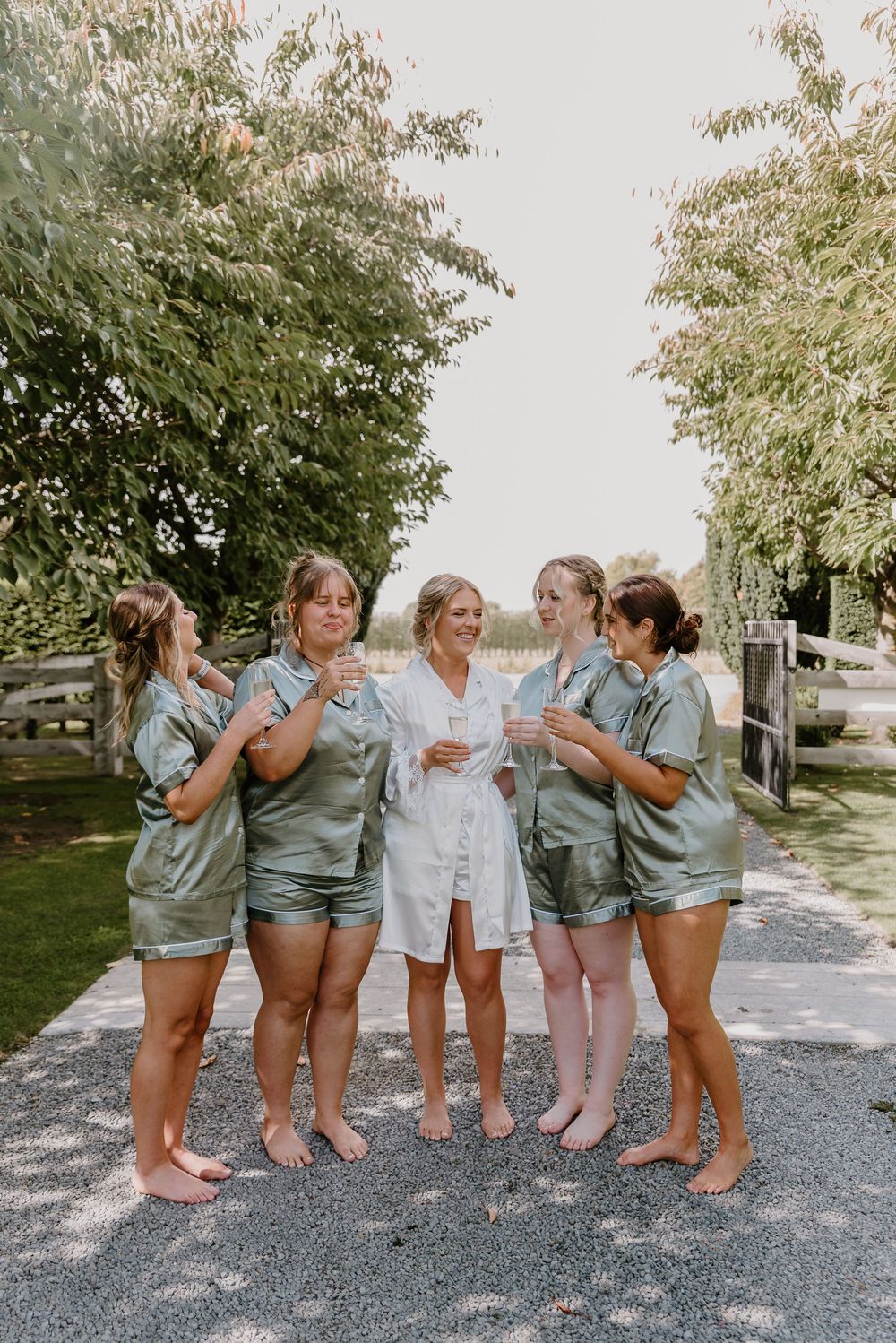 A group in matching sage green satin pajama sets gather outdoors while one person wears white on a sunny day.