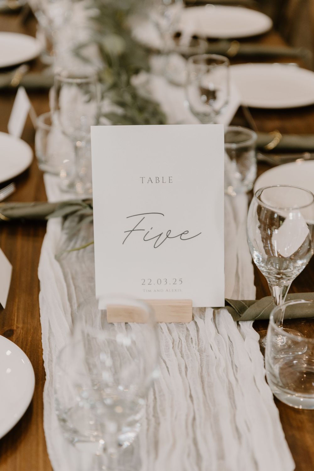 Elegant wedding table setting with white table number five card, glassware, and flowing fabric runner on wooden table.