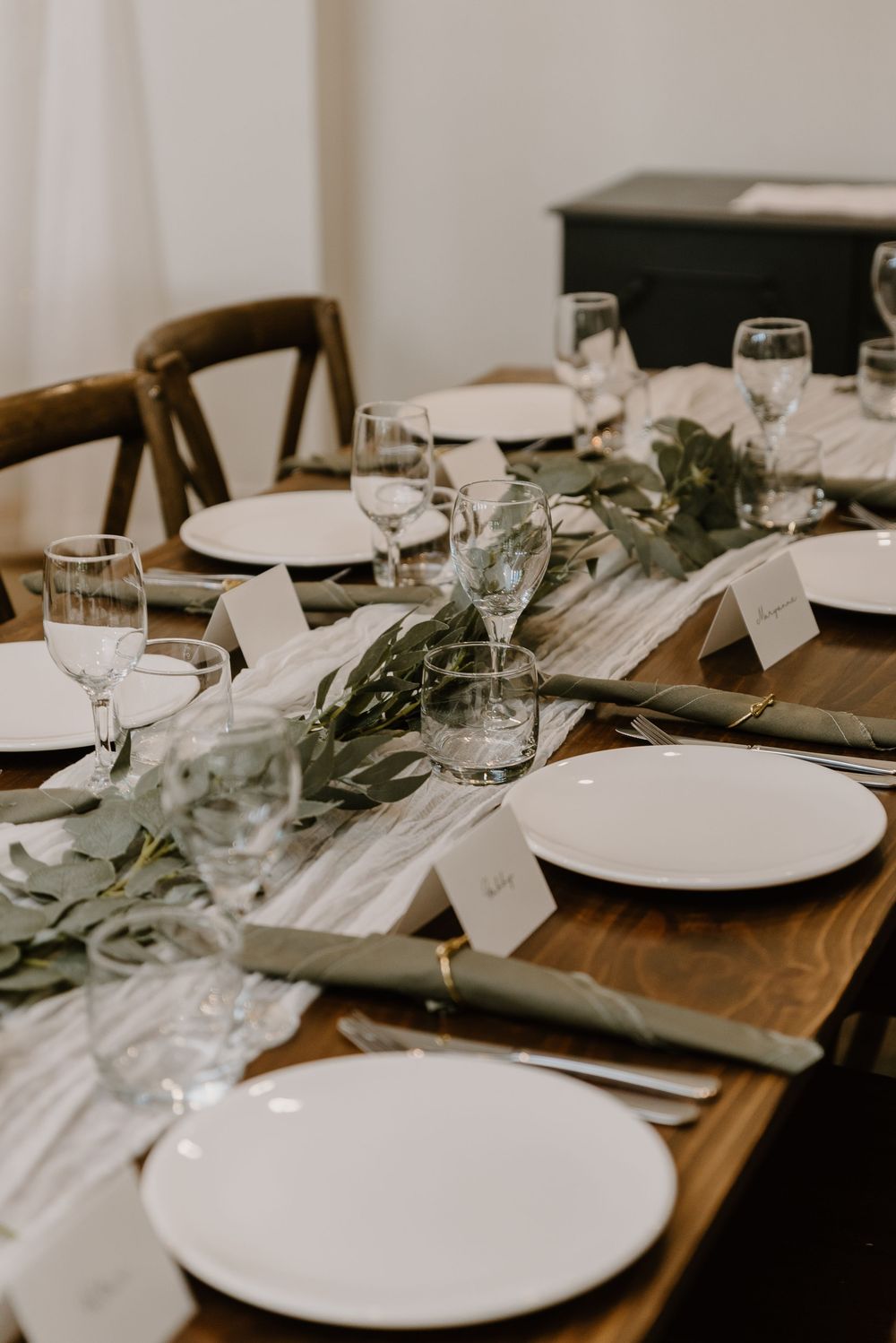 An elegant table setting with white plates, wine glasses, and eucalyptus garland on a rustic wooden table.