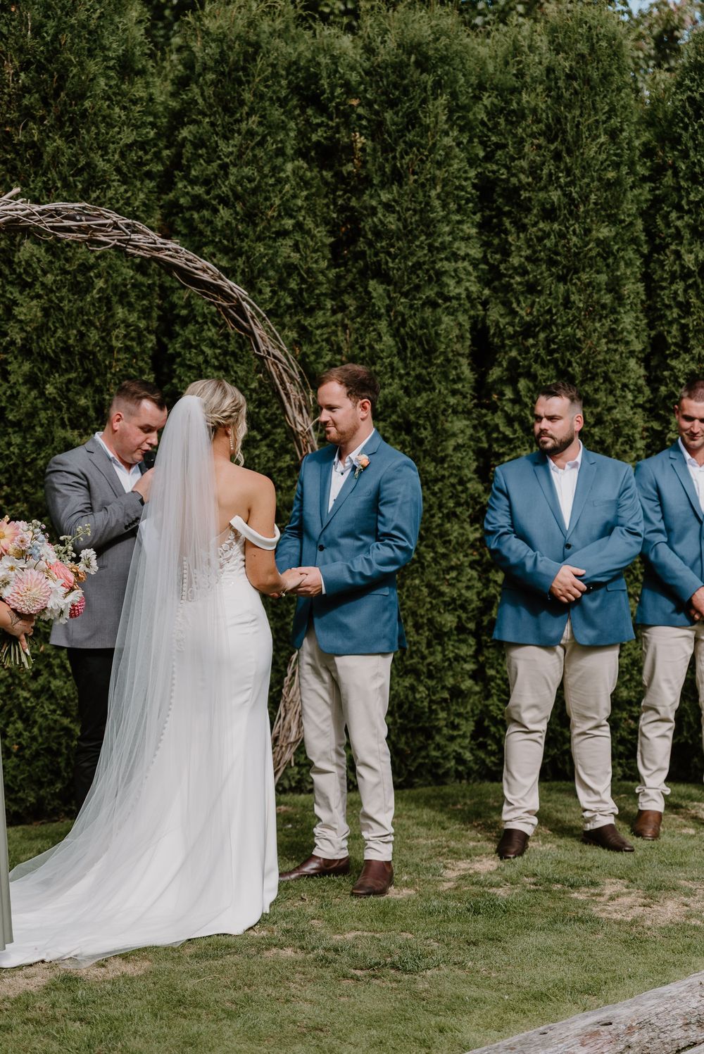 Wedding party poses outdoors against green hedges with groomsmen in blue suits and khaki pants.