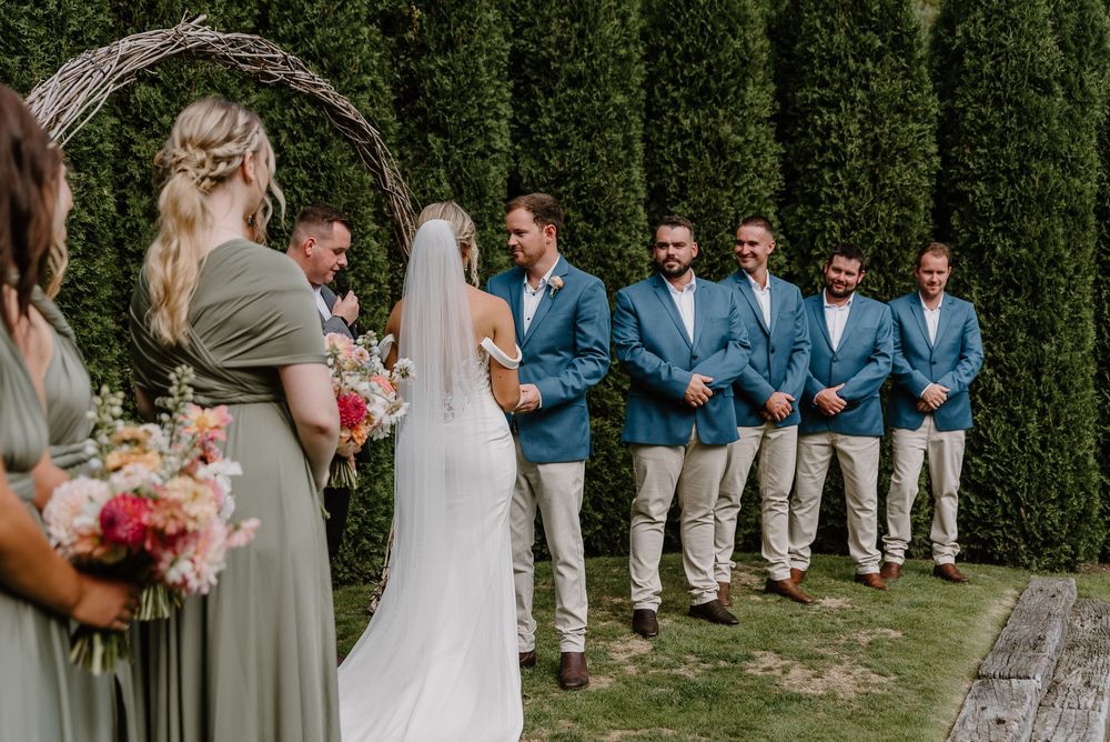 Elegant outdoor wedding party with bridesmaids in sage green dresses and groomsmen in blue suits posing together.
