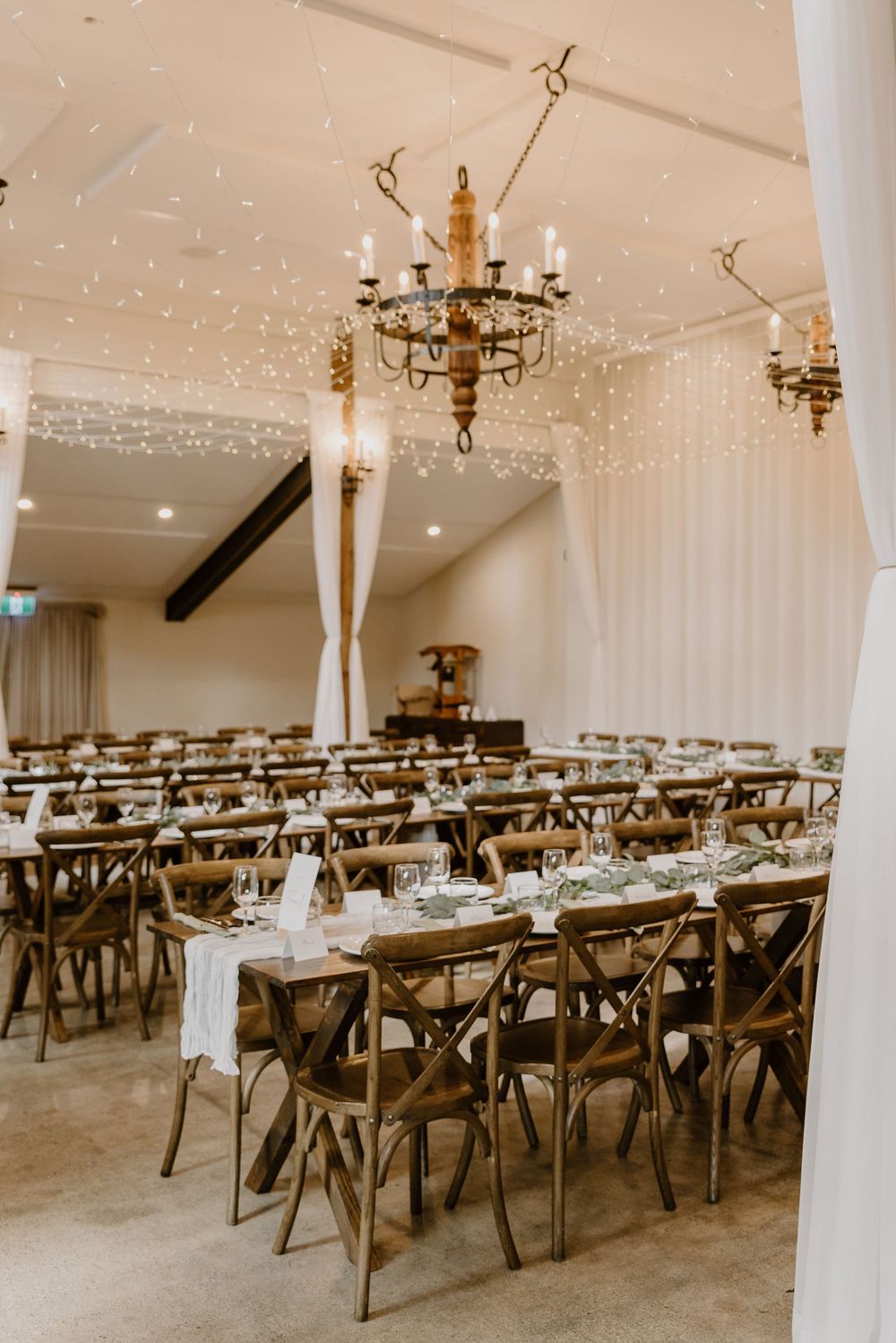 Elegant wedding reception venue with rustic wooden tables, brass chandeliers, and white drape decor illuminated by string lights.