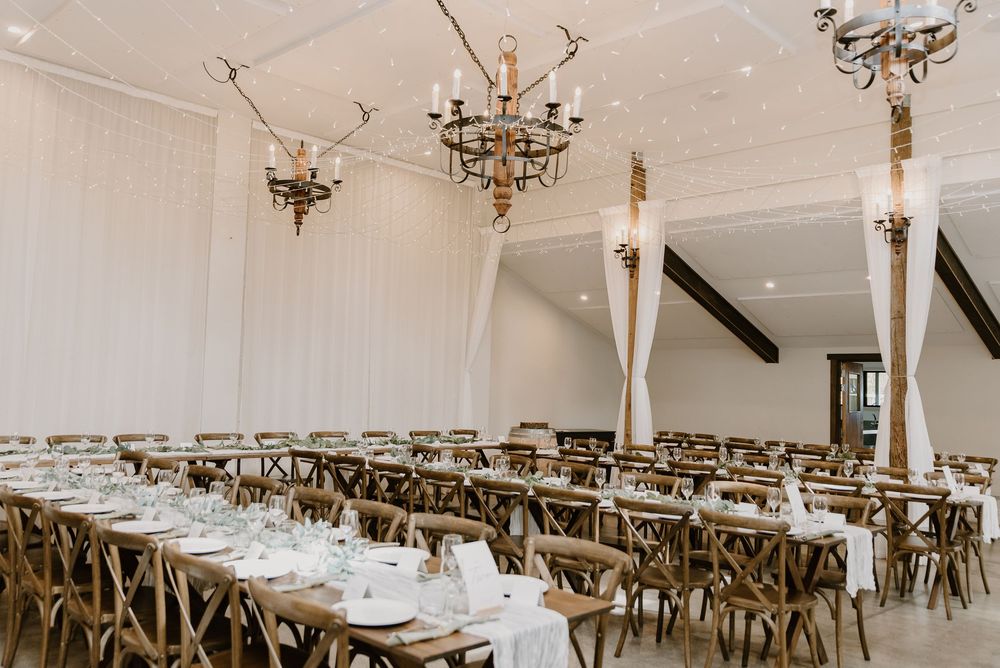 An elegant white reception venue with rustic cross-back chairs and brass chandeliers arranged for a formal dinner setting.