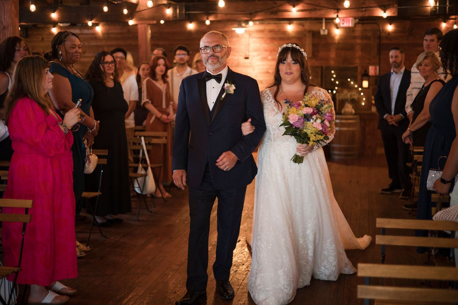 Wedding guests look on as the bride walks down the aisle in a rustic wooden barn venue illuminated by warm lighting.