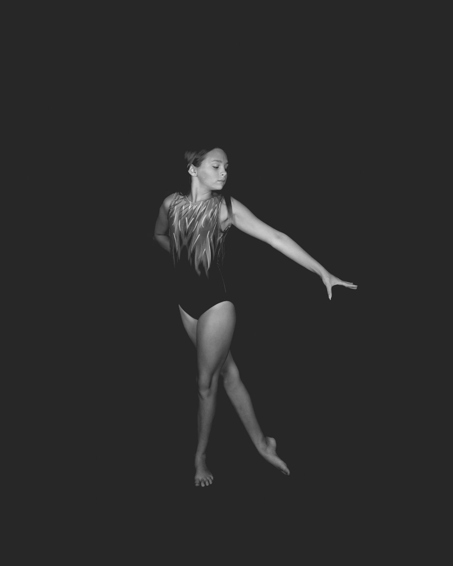 Graceful dancer in black leotard performs elegant pose against dark background in black and white photograph.