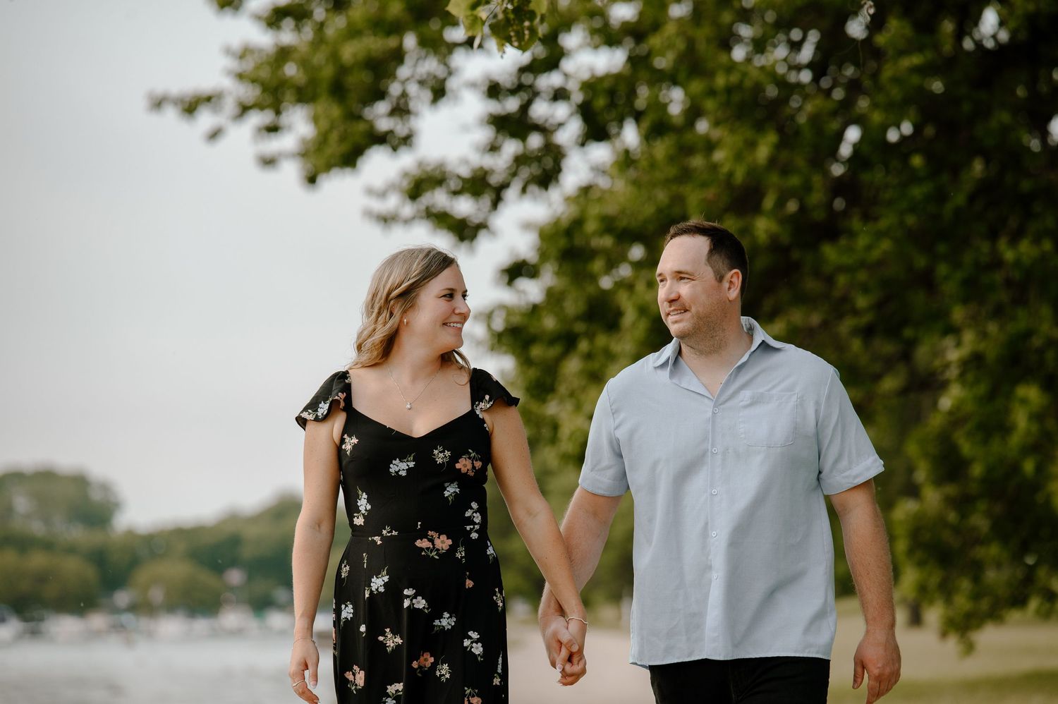 A couple walking hand in hand along a waterfront path with trees in the background on a summer evening.