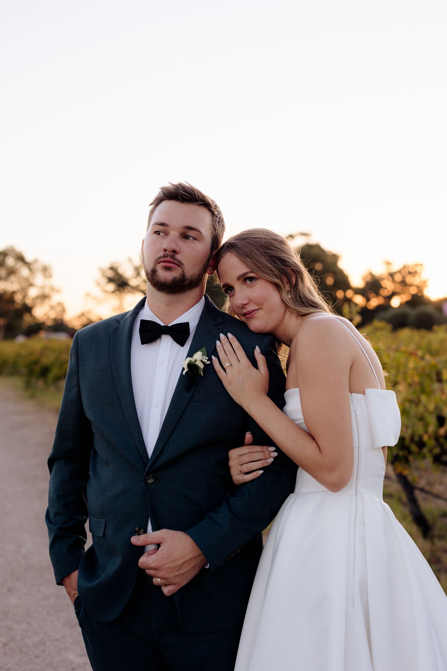 A couple in wedding attire pose together at sunset in a vineyard setting.