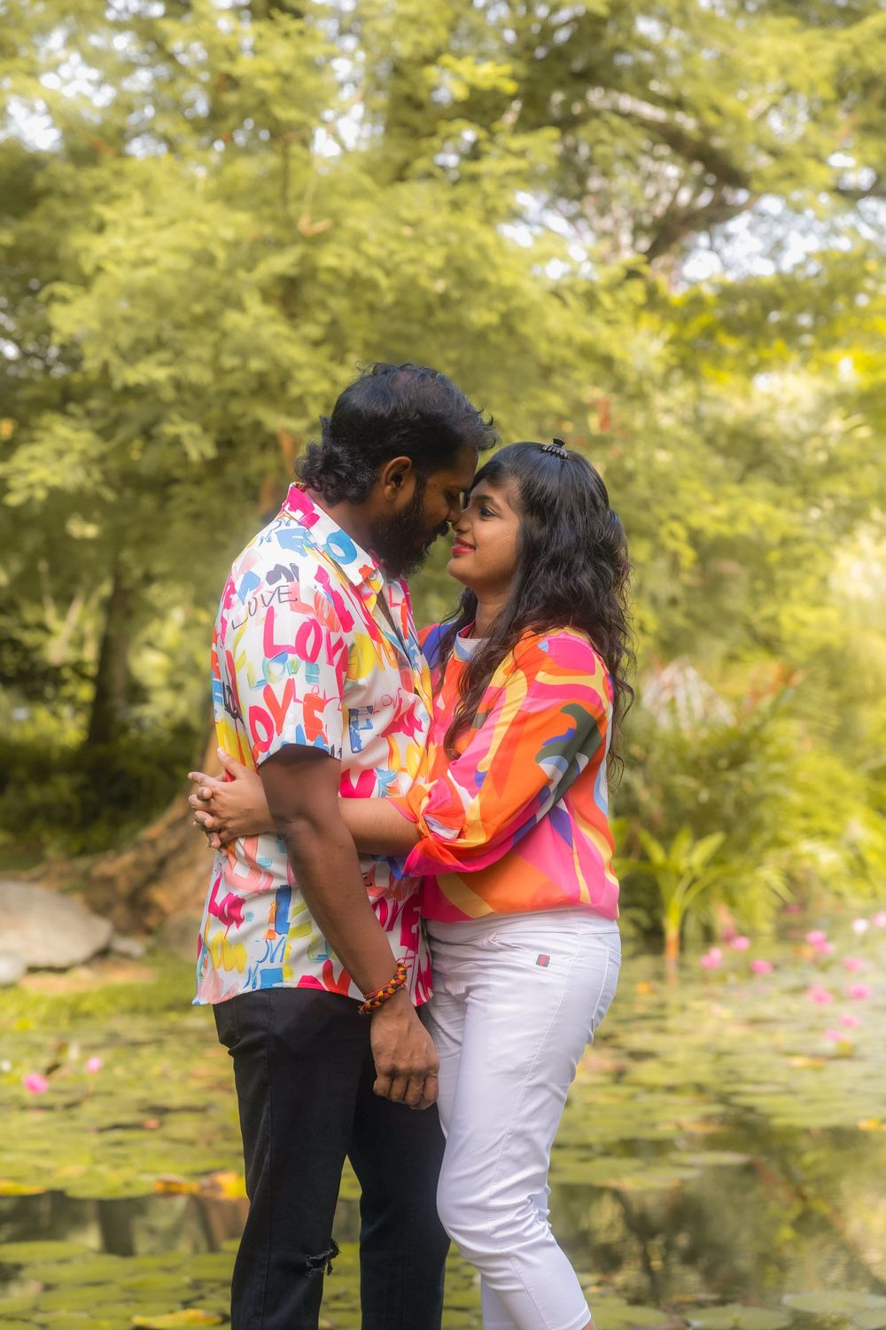 A romantic couple embraces in a sunlit park with lush green trees and foliage in the background.