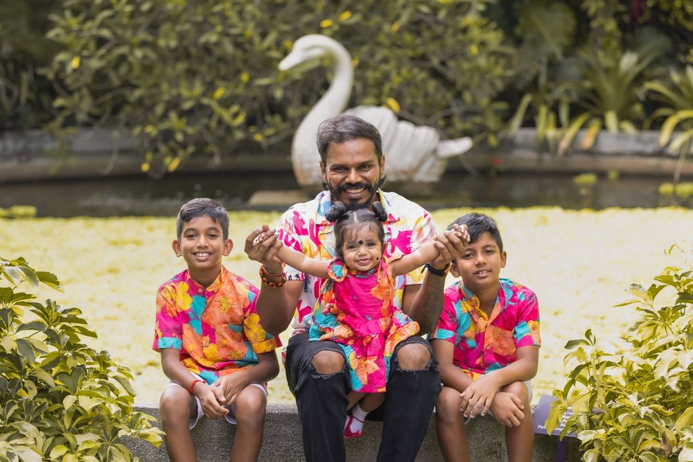 A group in colorful floral shirts sits on a concrete ledge by a pond with a white swan in the background.
