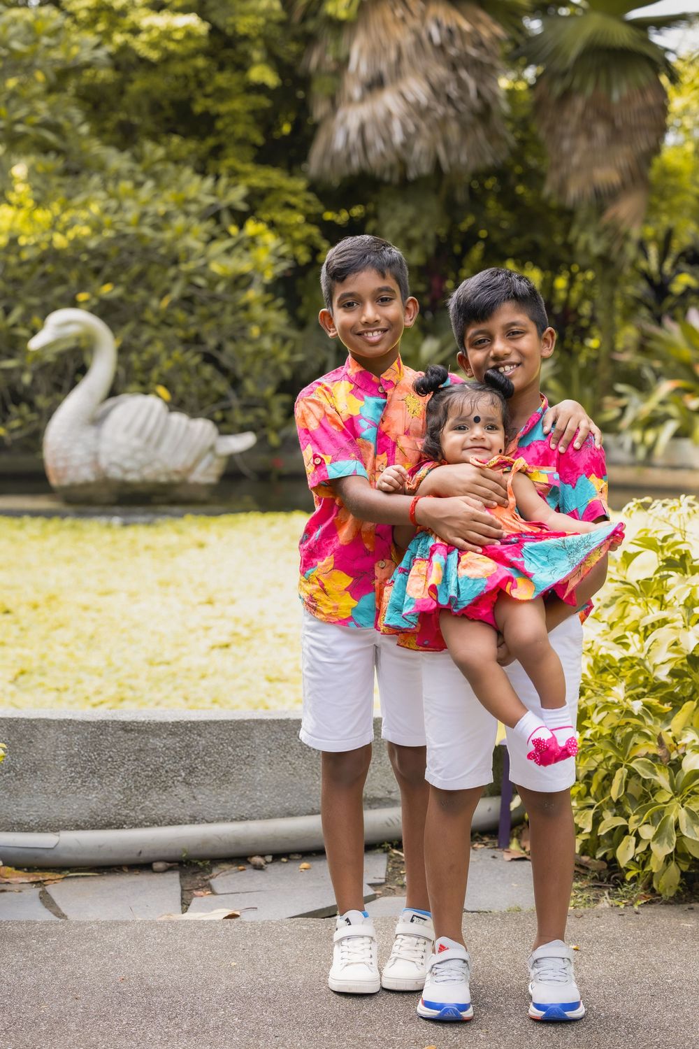 Three young siblings in colorful tie-dye shirts and white shorts pose together on garden steps with bright foliage.