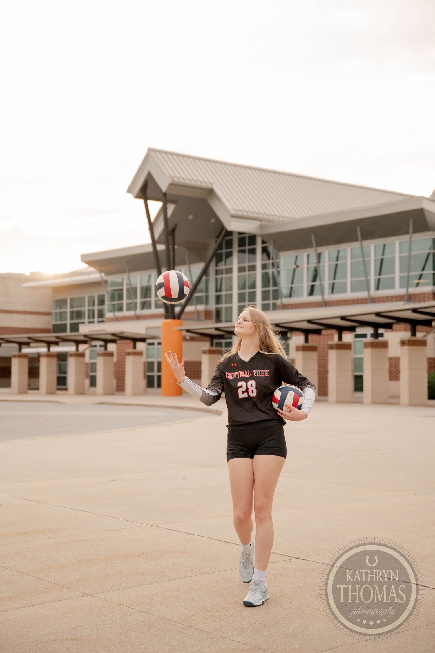 High school volleyball player tosses ball in front of modern school building during golden hour photoshoot.