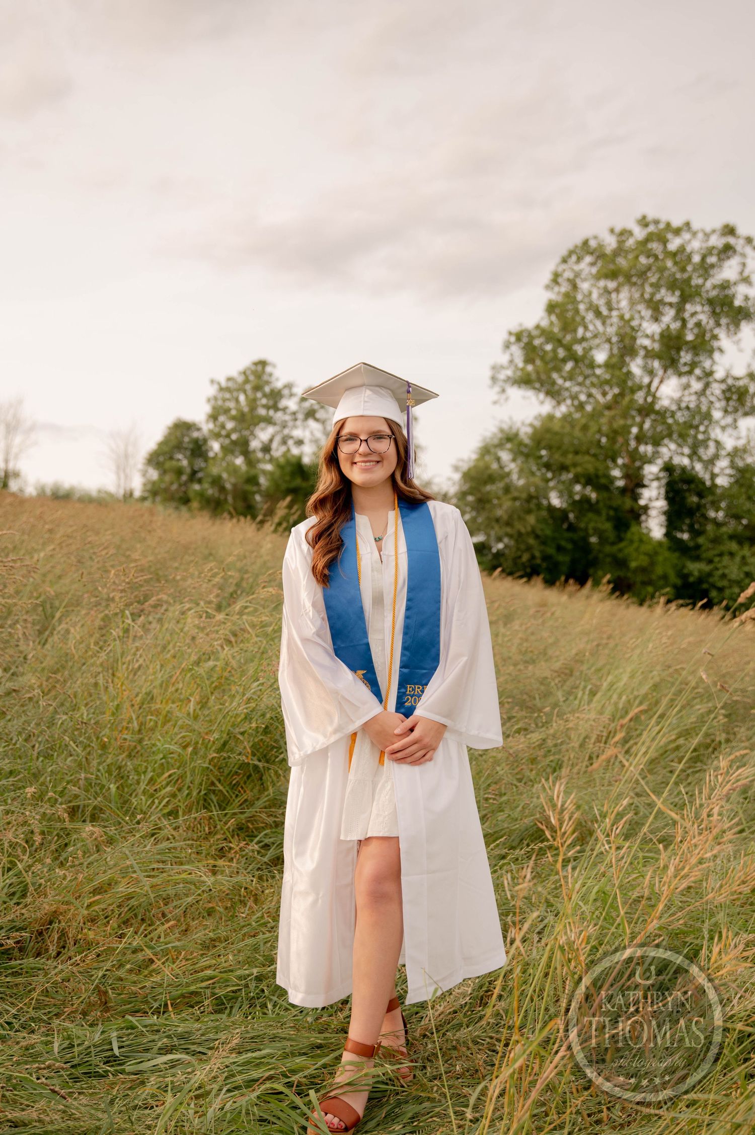 Graduate in white cap and gown with blue stole stands in tall grass field during golden hour photography session.
