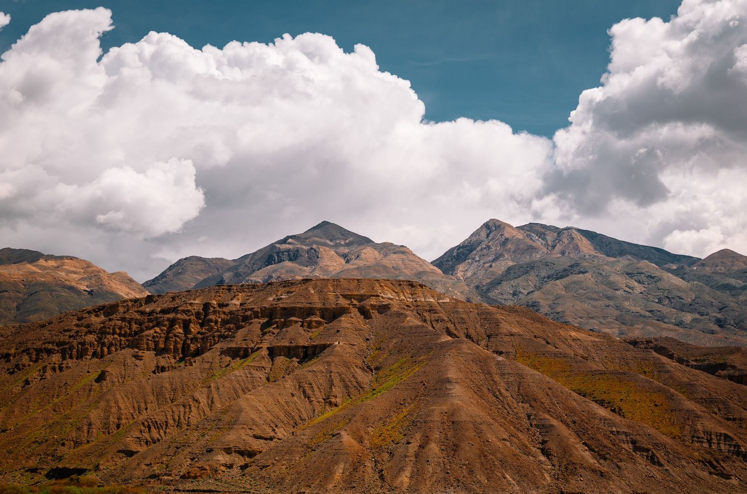 Rugged brown mountain peaks with eroded cliffs under billowing white clouds and blue sky.