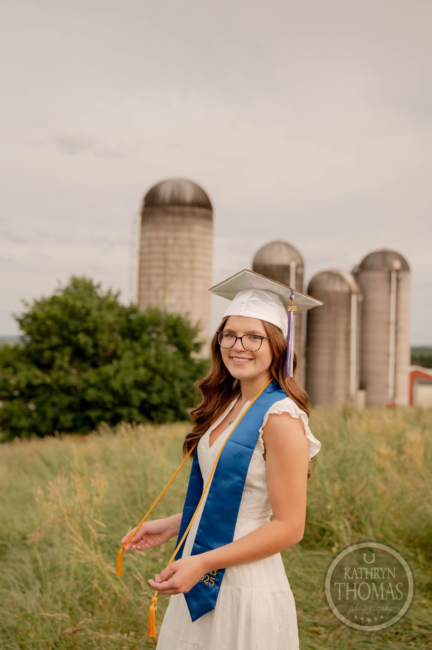 A graduate in white dress and blue sash poses in a field with tall agricultural silos in the background.