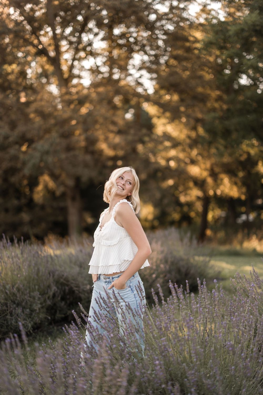 A person poses in a white sleeveless top and flowing skirt among blooming lavender at golden hour in a garden setting.