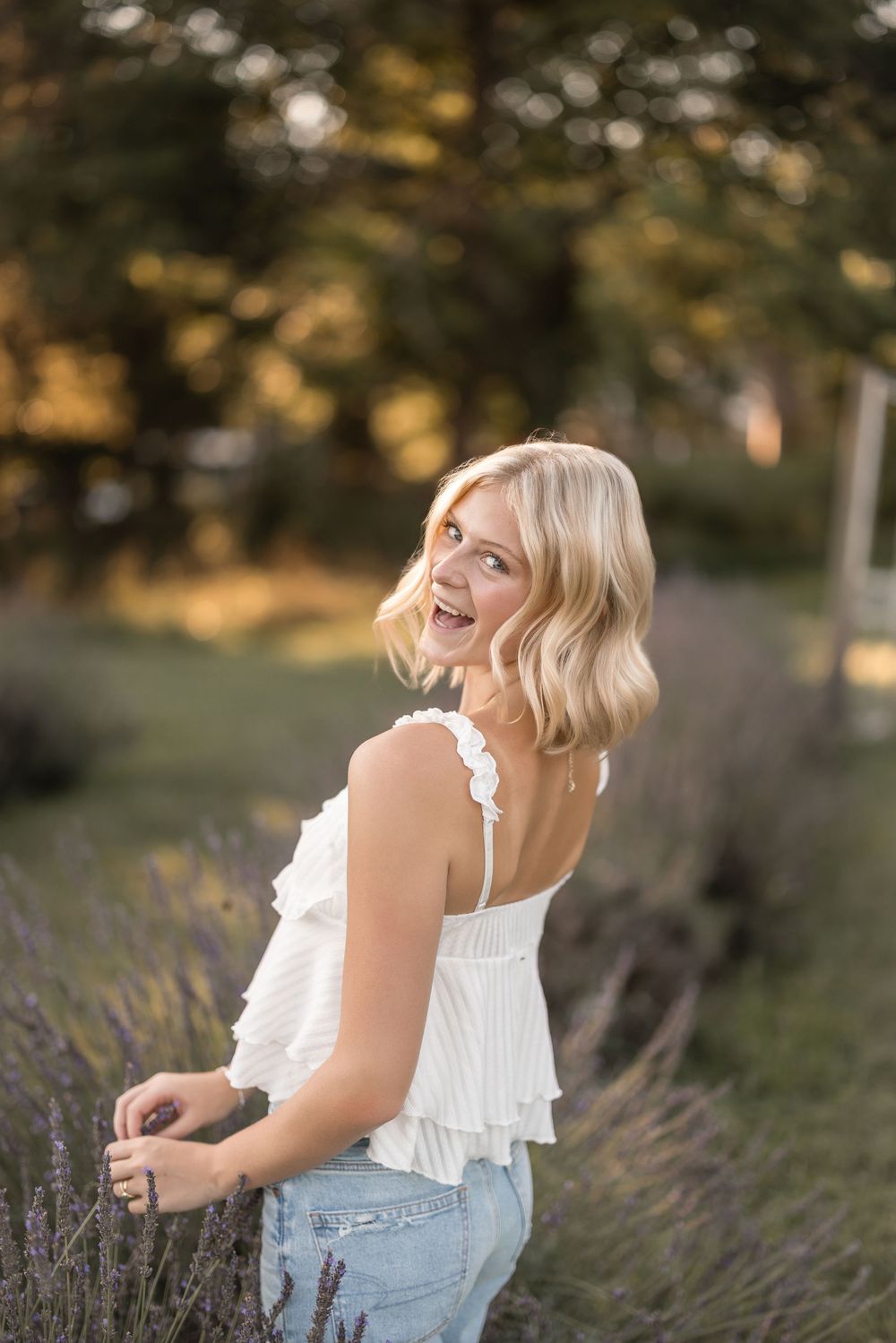 A happy person in a white ruffled top sits among blooming lavender flowers in a sunny outdoor garden.