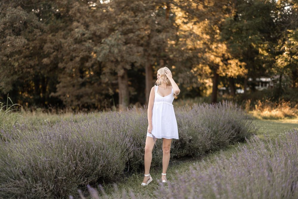 A person in a white summer dress stands among blooming lavender flowers during golden hour at sunset.