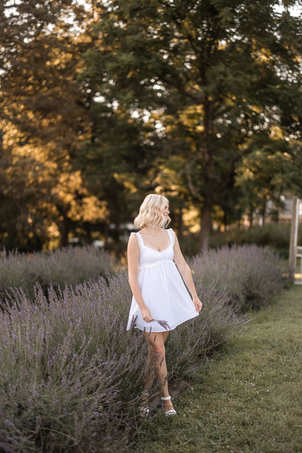 Woman in white sundress stands in a lavender field at golden hour with sunlit trees in the background.