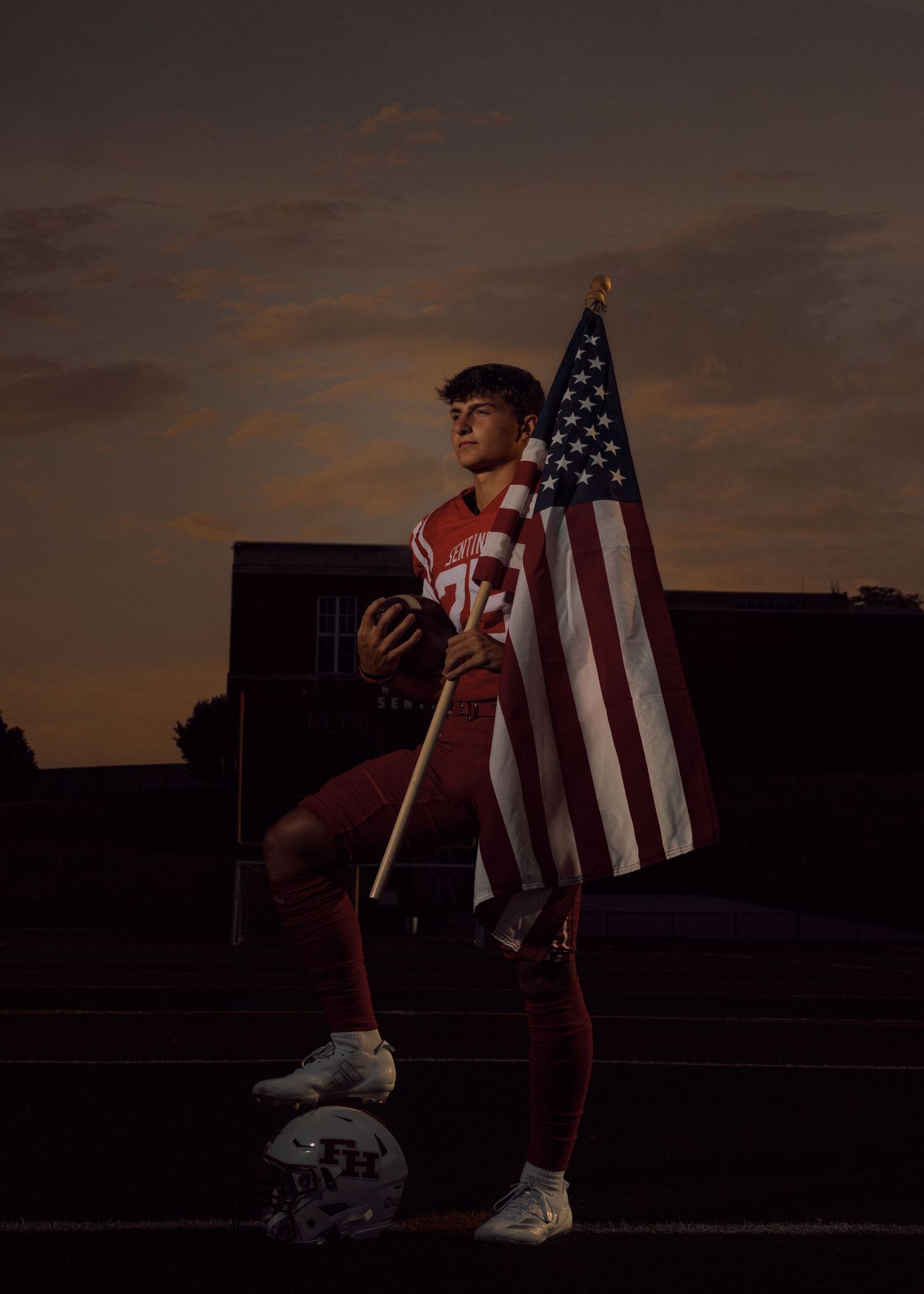 Silhouetted figure holds American flag against dramatic sunset sky in urban setting.